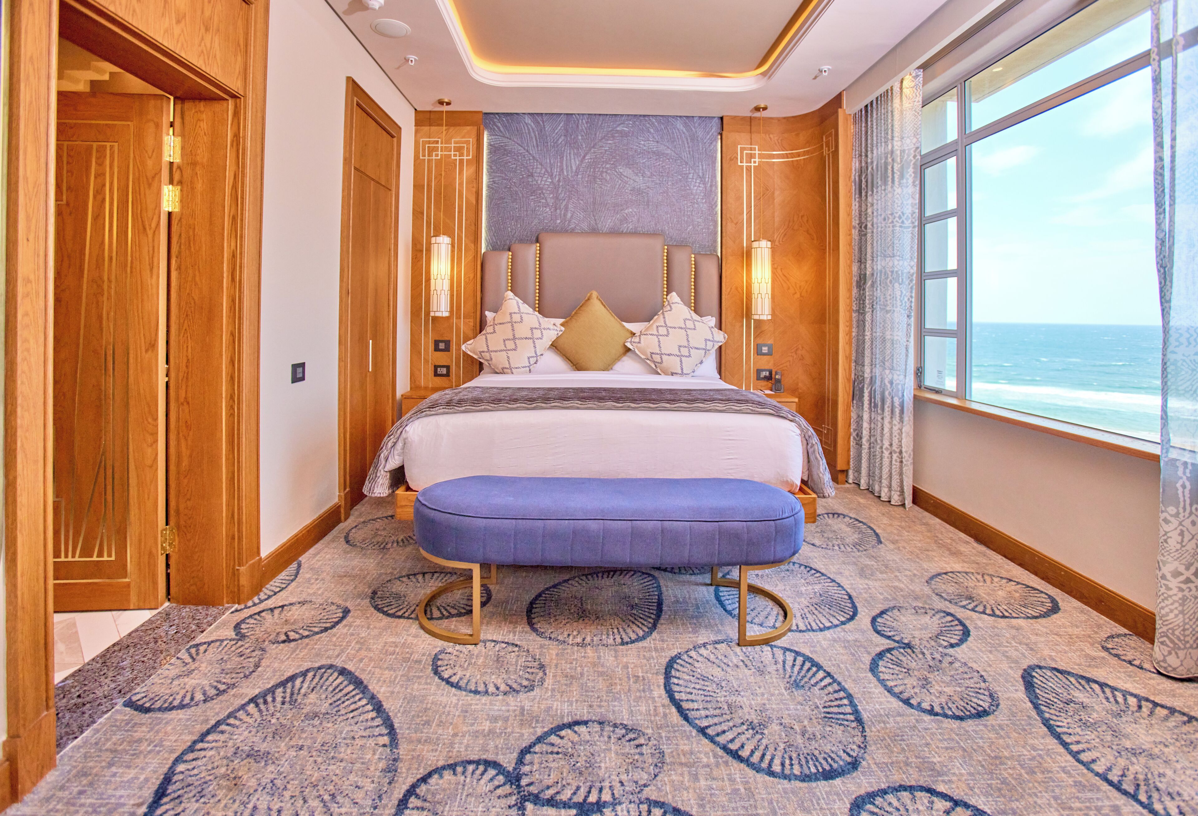A bedroom with a large bed, a bench, a view of the ocean, and a patterned carpet.