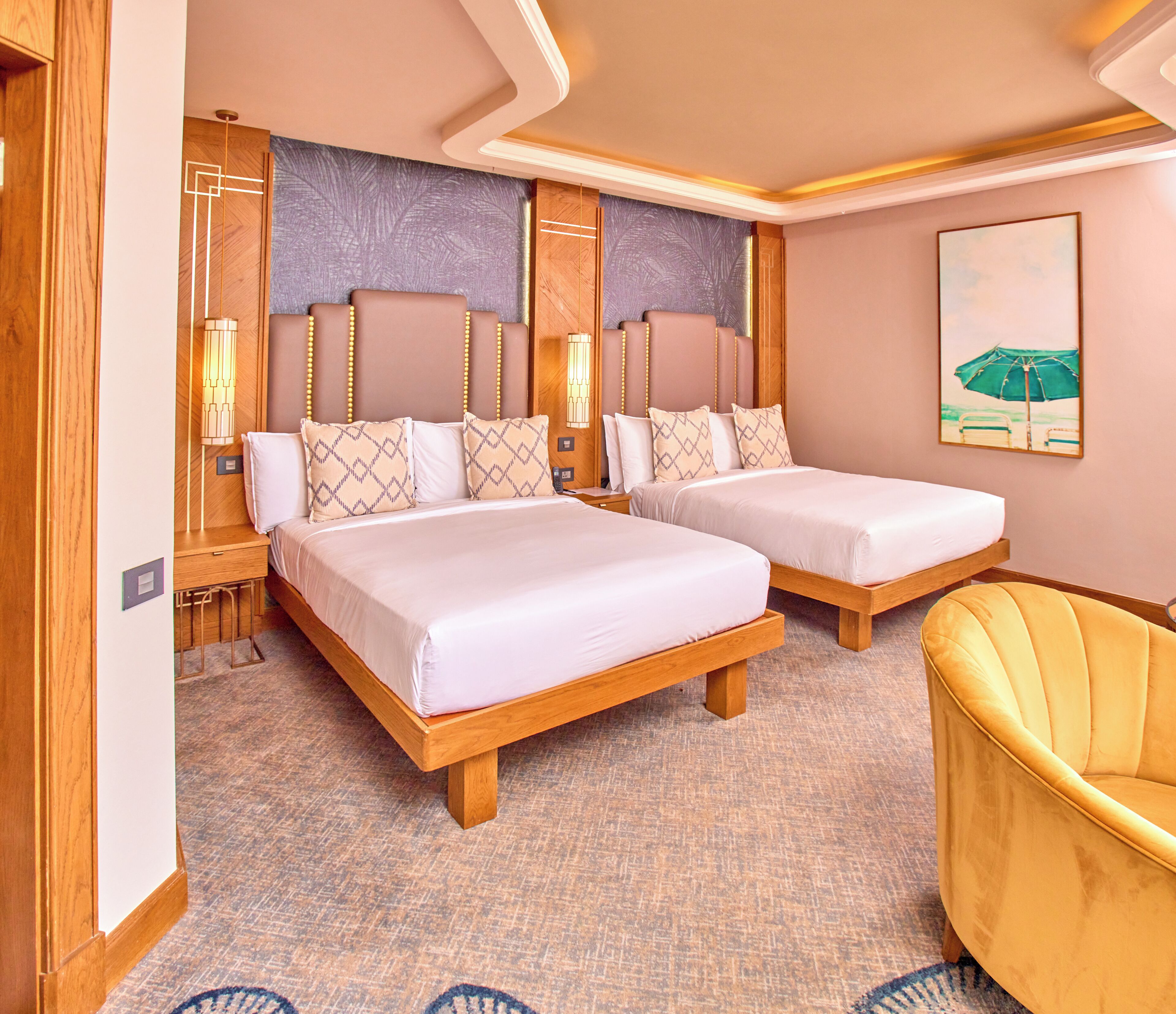 A hotel room with two beds, wooden headboards, a yellow armchair, and a painting of a beach scene.