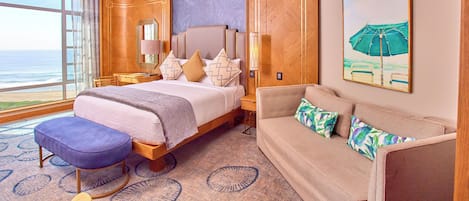 A hotel room with a large bed, a sofa, a small table, and a view of the ocean.