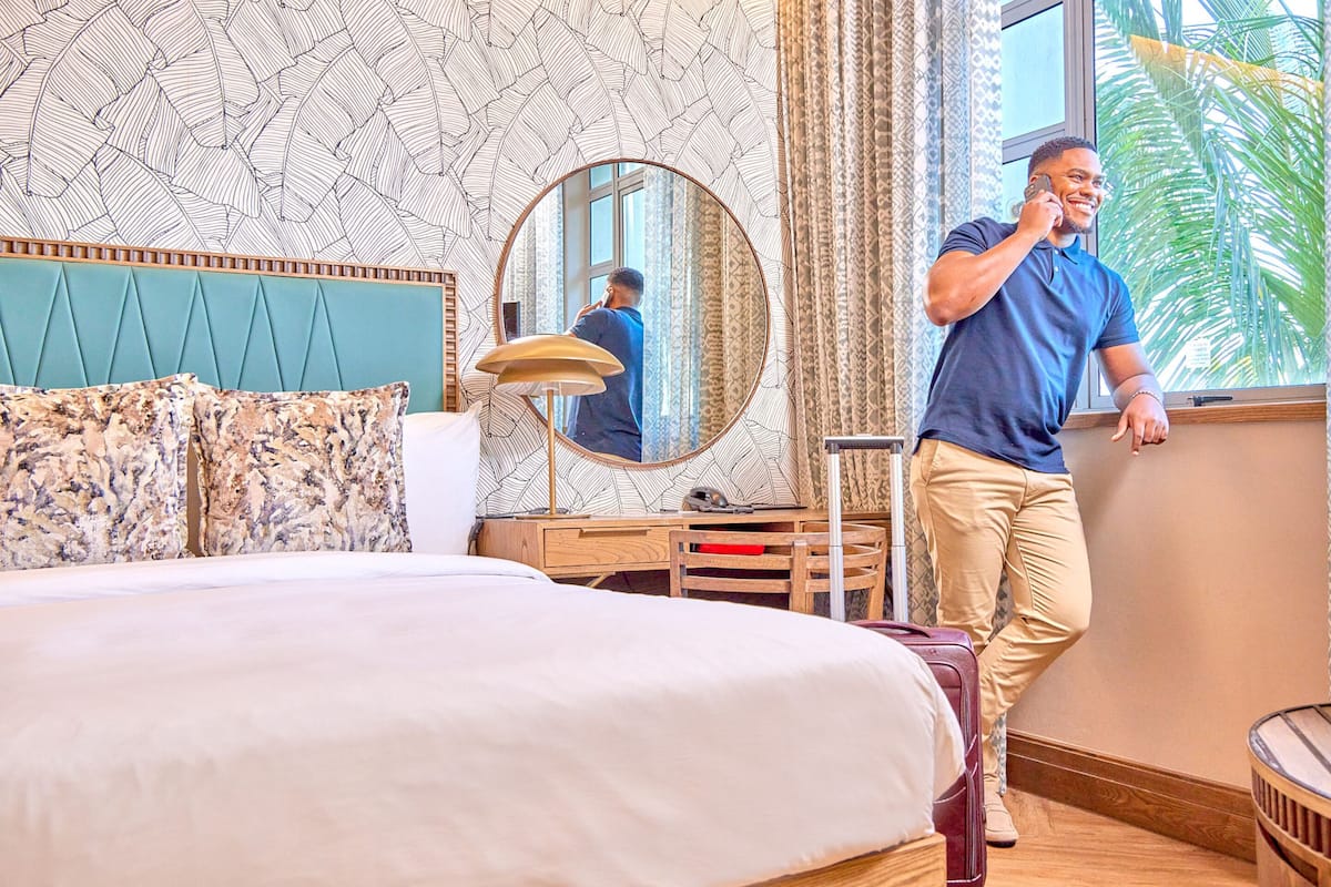 Habitación de hotel con una cama grande, un escritorio con lámpara, un espejo y una ventana que da a vistas de palmeras.