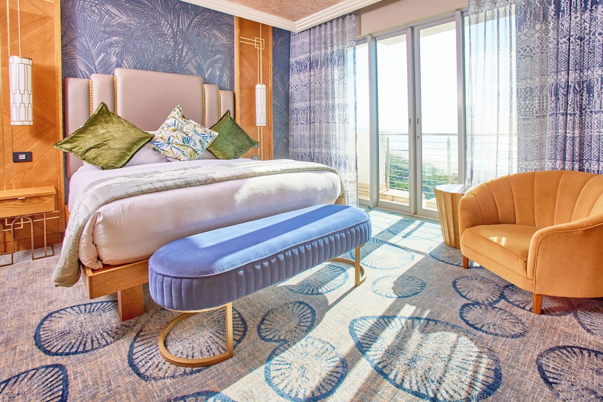 Una habitación de hotel con cama, un banco, una silla, un ventanal grande y una alfombra estampada.
