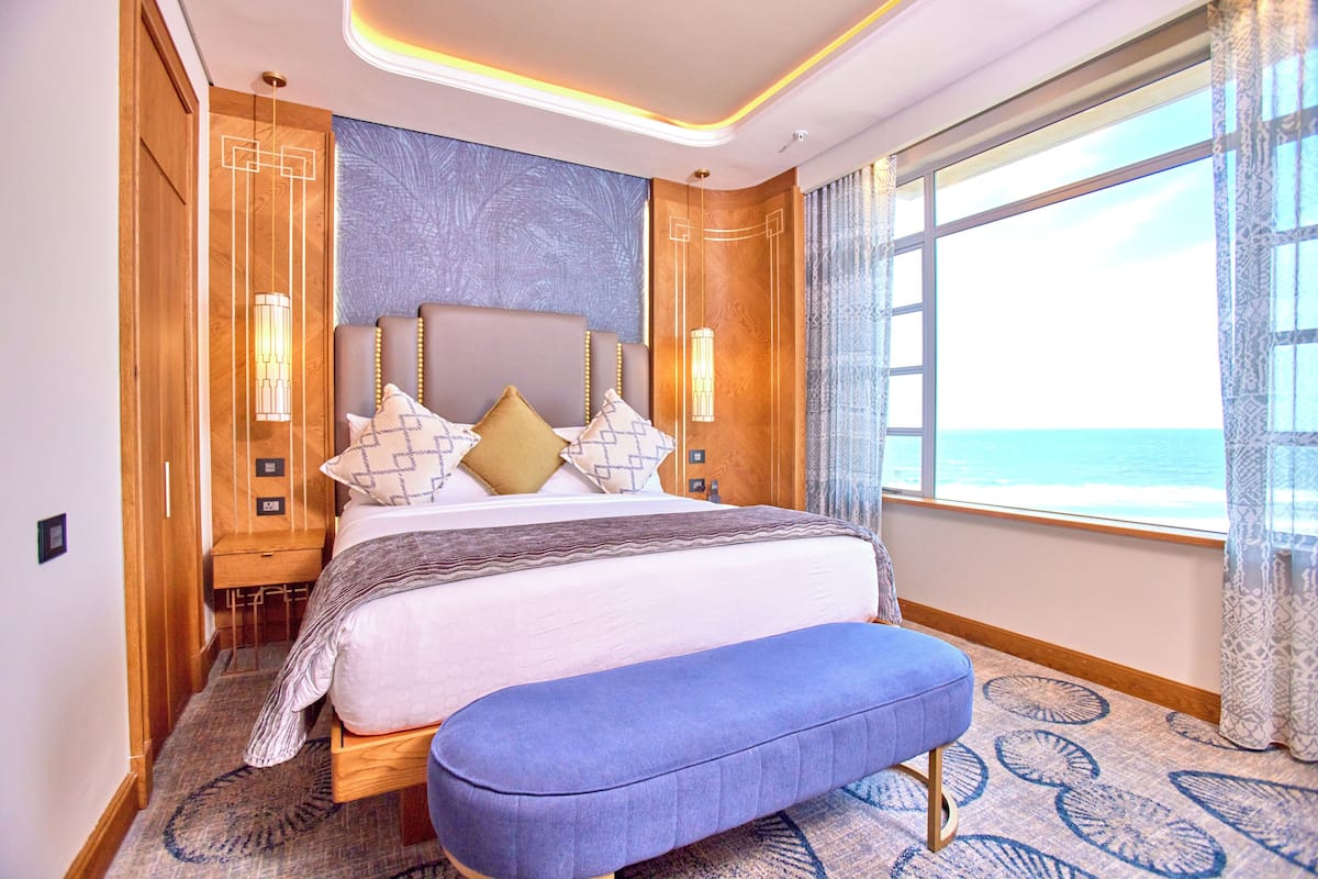 Un dormitorio con una cama grande, un banco, una mesita de noche y vistas al mar.