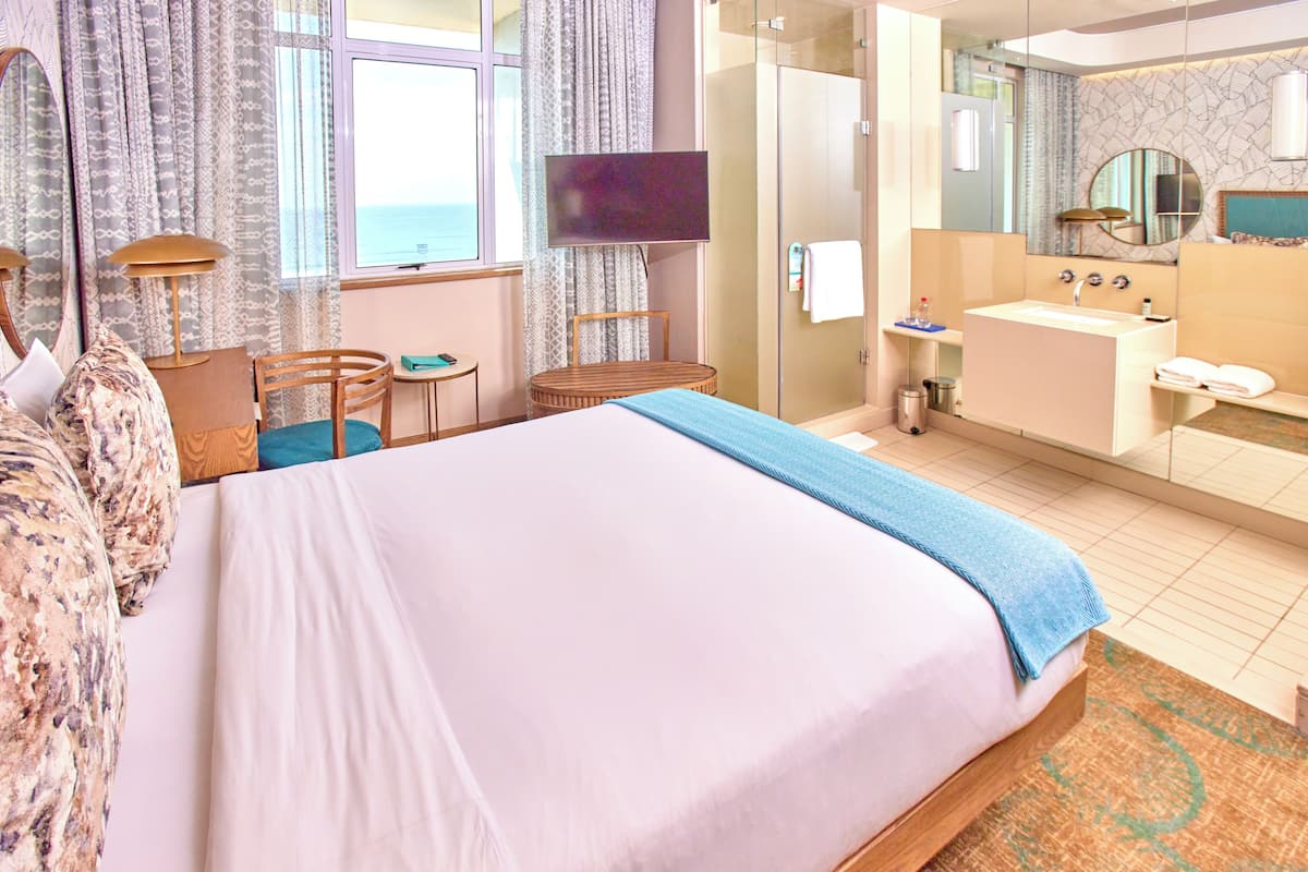 Habitación de hotel con cama, escritorio, silla, televisor, baño con lavabo y espejo, y vista al mar por la ventana.