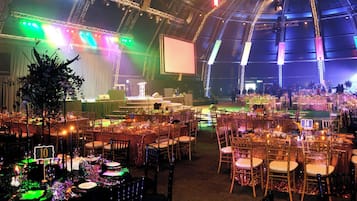 Un amplio espacio interior con varias mesas redondas y sillas, un escenario con una pantalla grande, y una estructura parecida a una carpa con iluminación.