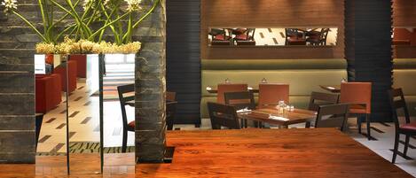 Een restaurant met een houten tafel en stoelen, een stenen pilaar met bloemen, en een zithoek met een balie.