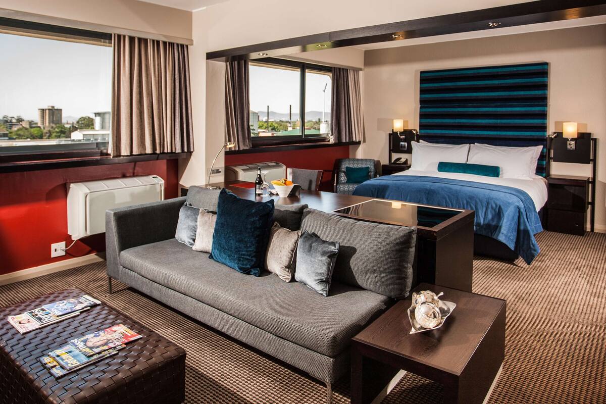 Ein Hotelzimmer mit Sofa, einem kleinen Tisch, einem Bett mit blauer Bettdecke, einem Nachttisch mit Lampe und Blick auf die Stadt durch große Fenster.