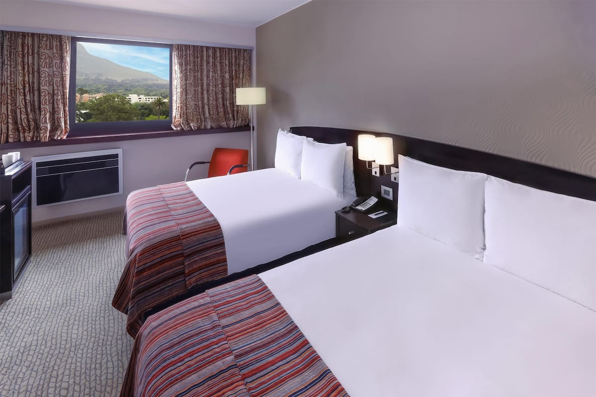 Ein Hotelzimmer mit zwei Betten, einem Schreibtisch, einem Stuhl, einer Lampe und Blick auf Berg und Stadt.
