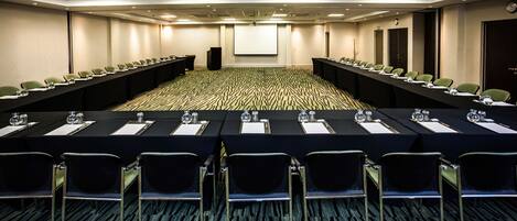 A conference room with a long table, rows of chairs, a projector screen, and a carpeted floor.