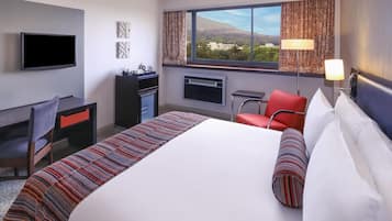 Habitación de hotel con una cama grande, un escritorio, una silla, un televisor y ventana con vista a edificios y árboles.