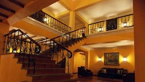 Un grand escalier orné de balustrades menant à un deuxième étage, un hall d’entrée avec un canapé et un tableau au mur, et un sol carrelé.