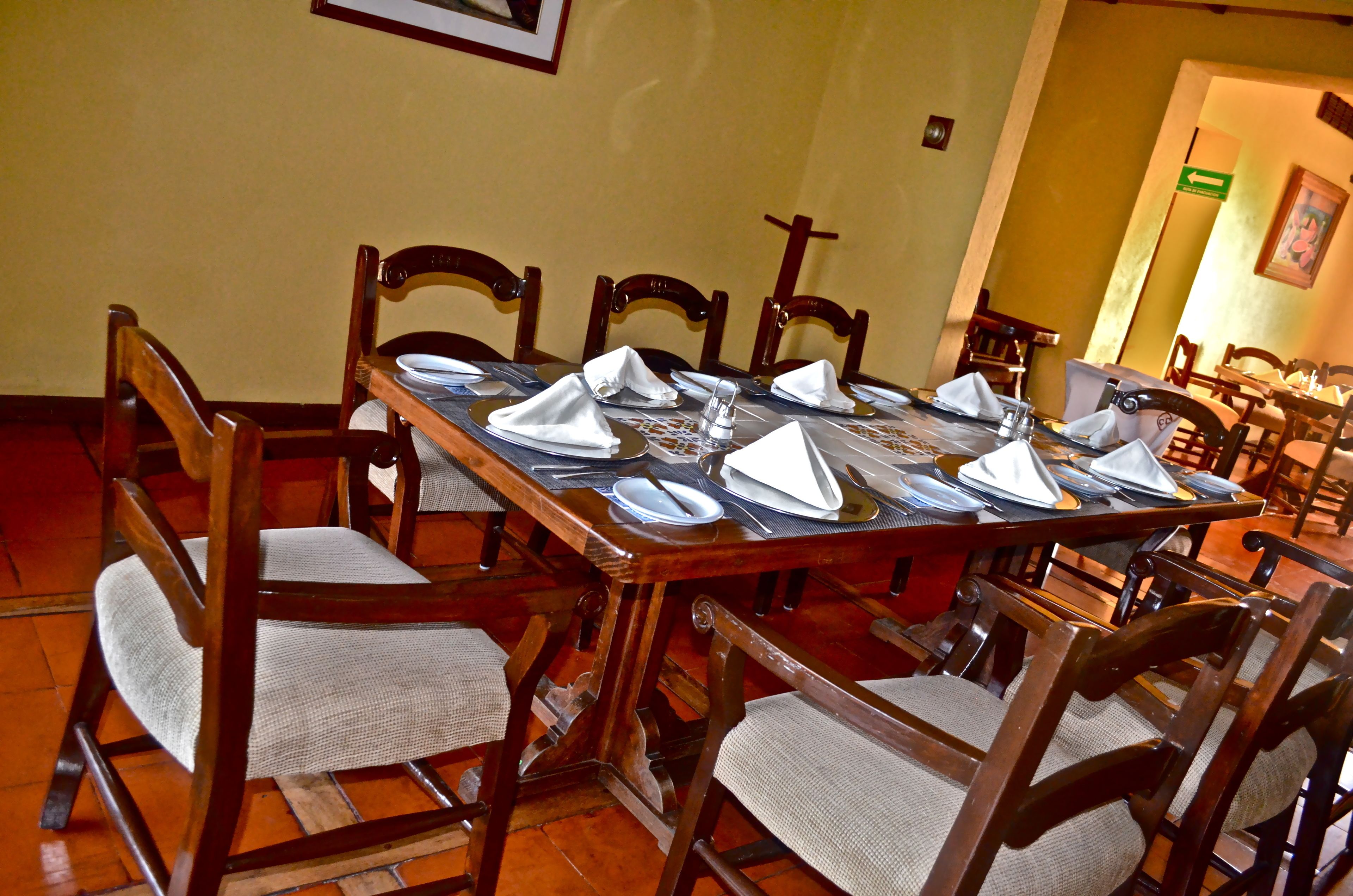 Une table à manger dressée pour six personnes, avec des chaises en bois, une nappe à motifs et un panneau de signalisation vert accroché au mur.