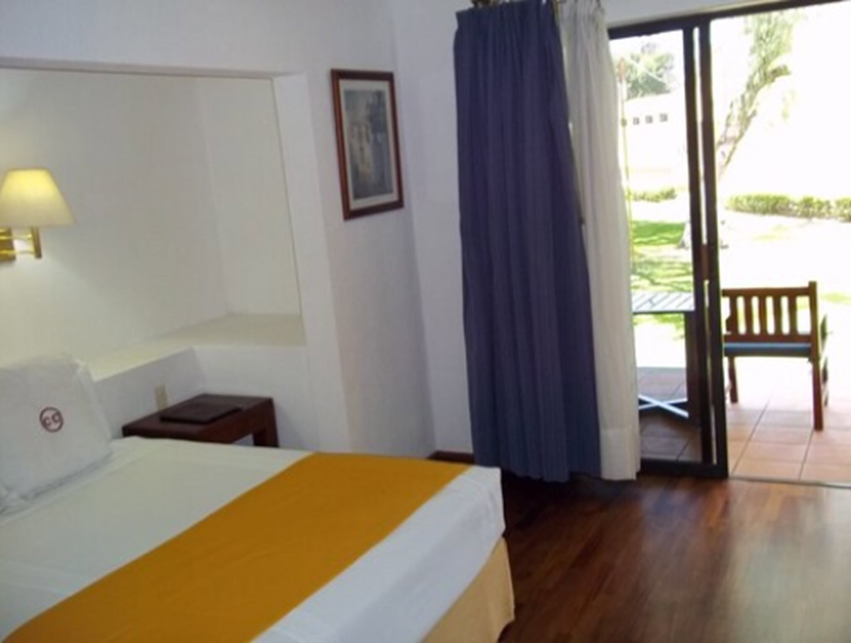 Habitación de hotel con cama, mesita de noche, lámpara, un cuadro en la pared y vistas al exterior a través de una puerta.
