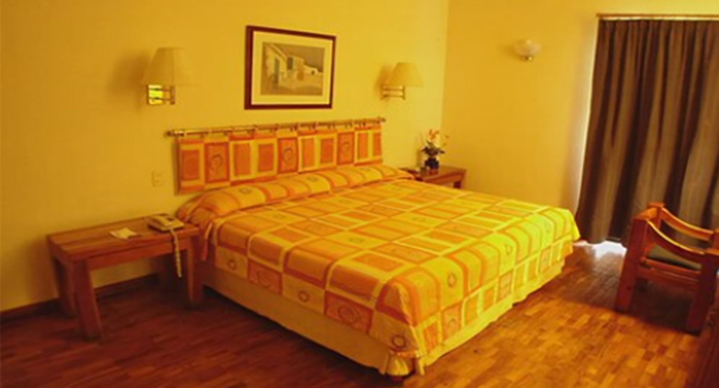 A hotel room with a large bed, wooden floor, a nightstand with a lamp, a chair, and a framed picture on the wall.