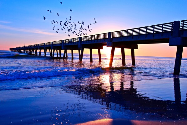 Um píer que se estende para o oceano ao pôr do sol, com pássaros voando acima.