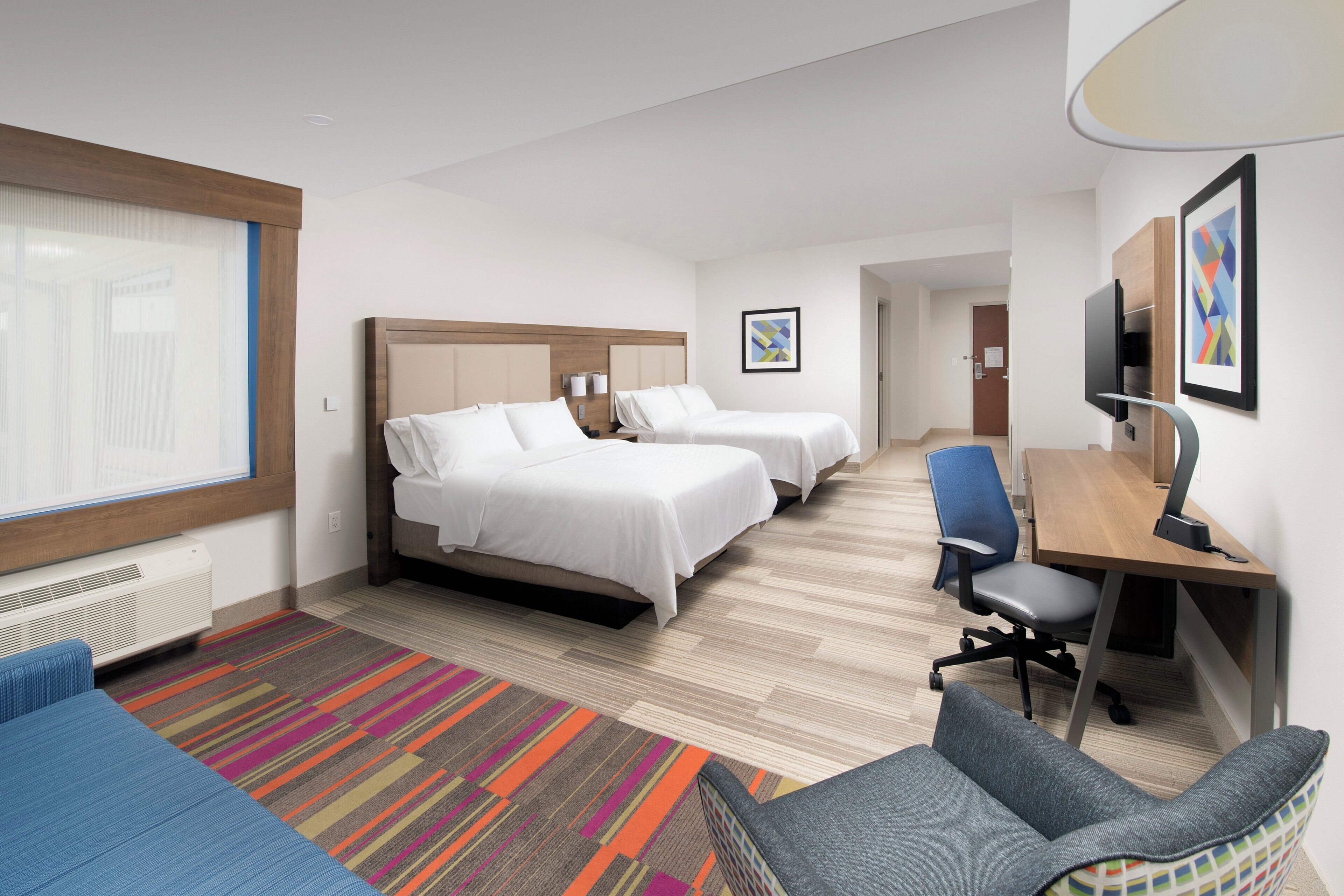 Habitación de hotel con dos camas, un escritorio, una silla, un televisor y una alfombra colorida.
