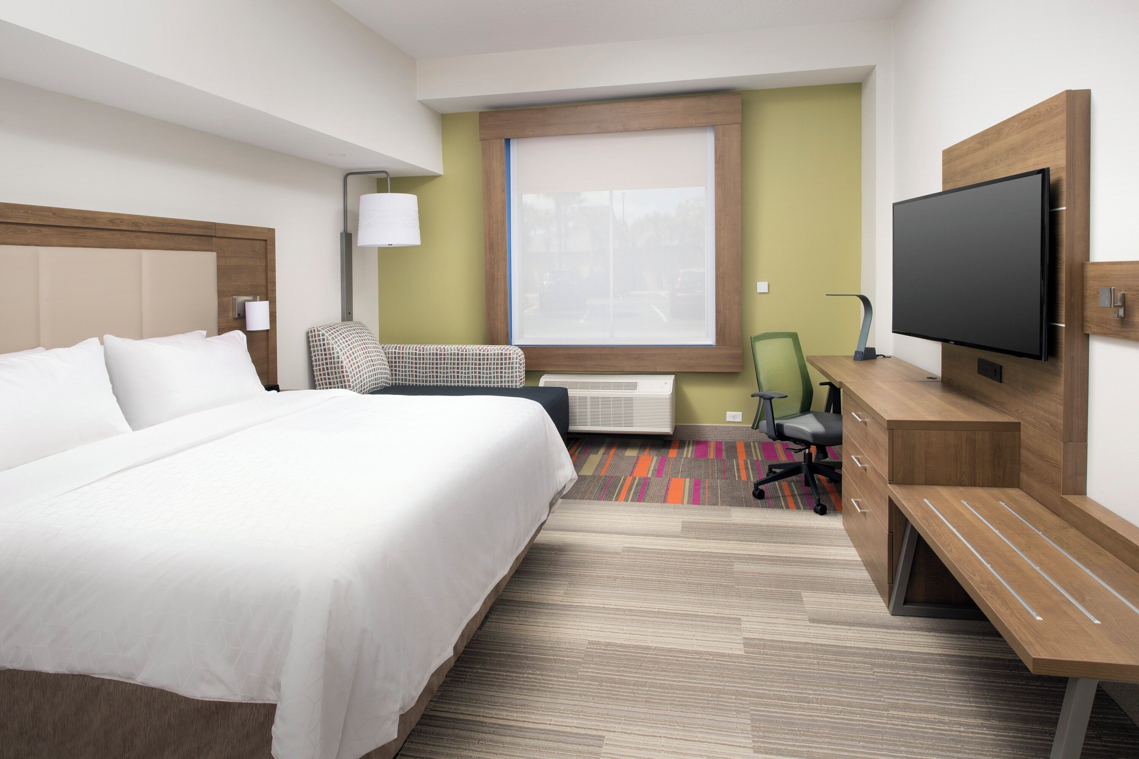 Quarto de hotel com cama, escrivaninha, cadeira, televisão e janela.