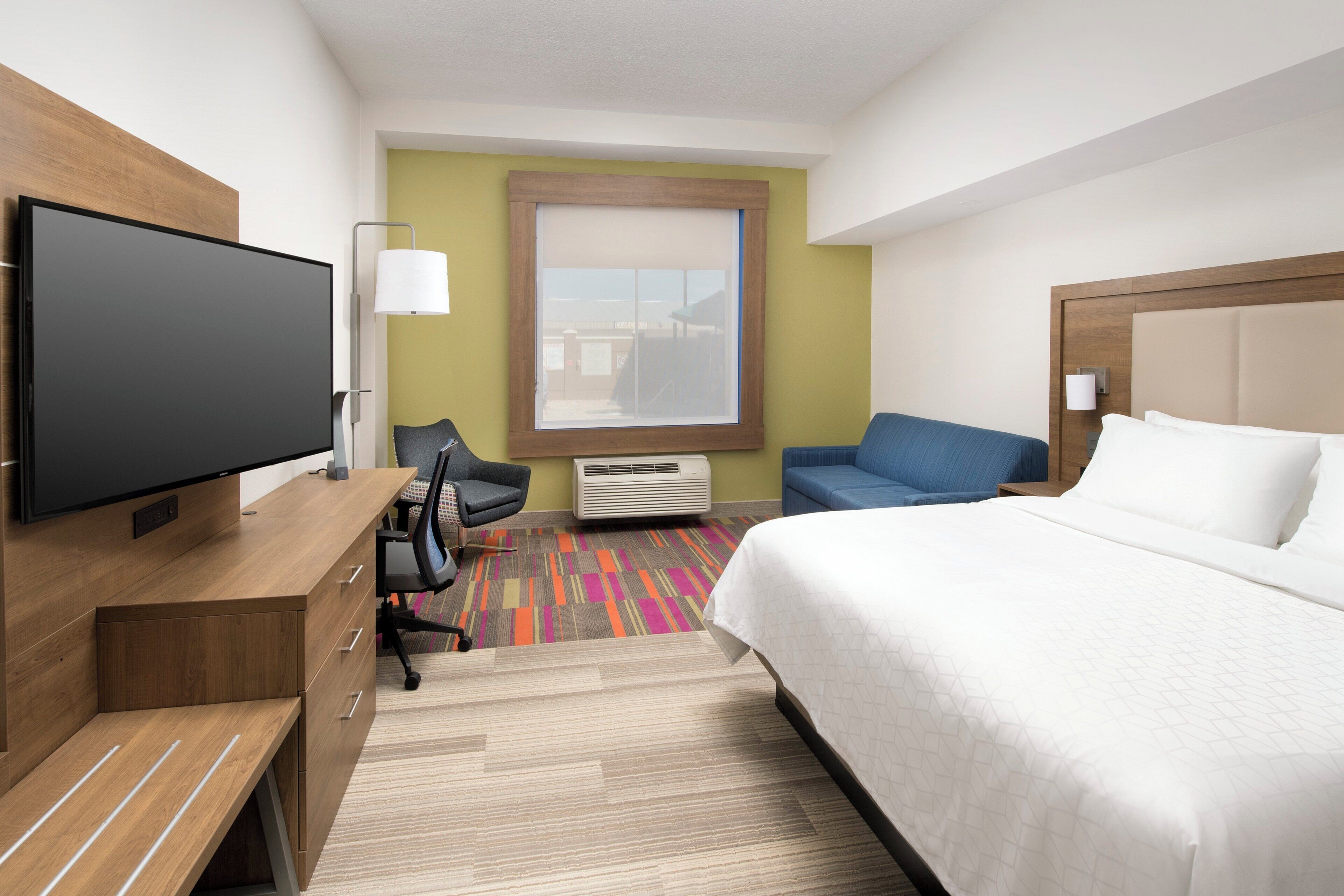 Une chambre d’hôtel comprenant un lit, un bureau avec un téléviseur à écran plat, une chaise, une fenêtre et une lampe fixée au mur.