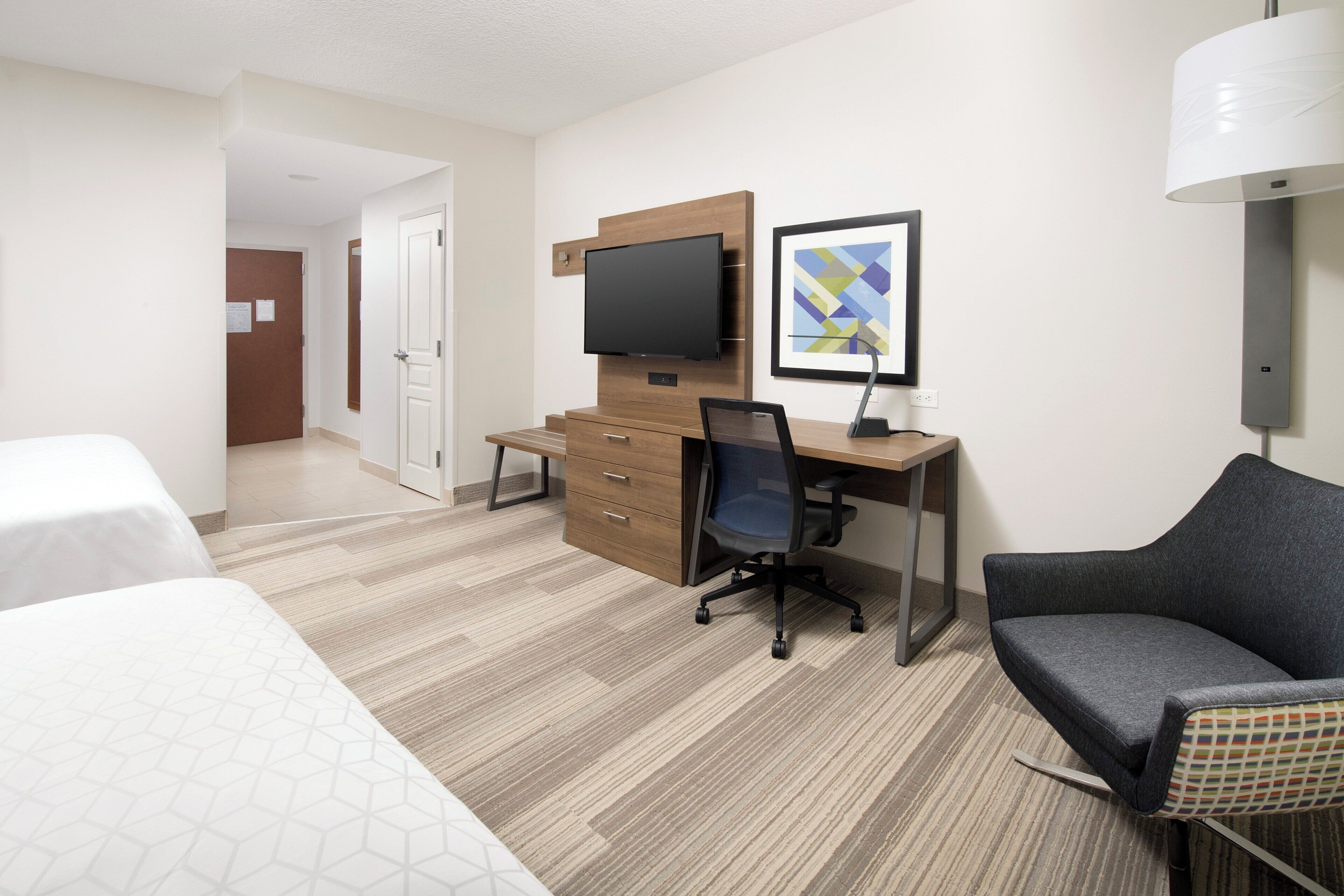 A hotel room with a bed, a desk, a chair, a television, and a wall-mounted lamp.