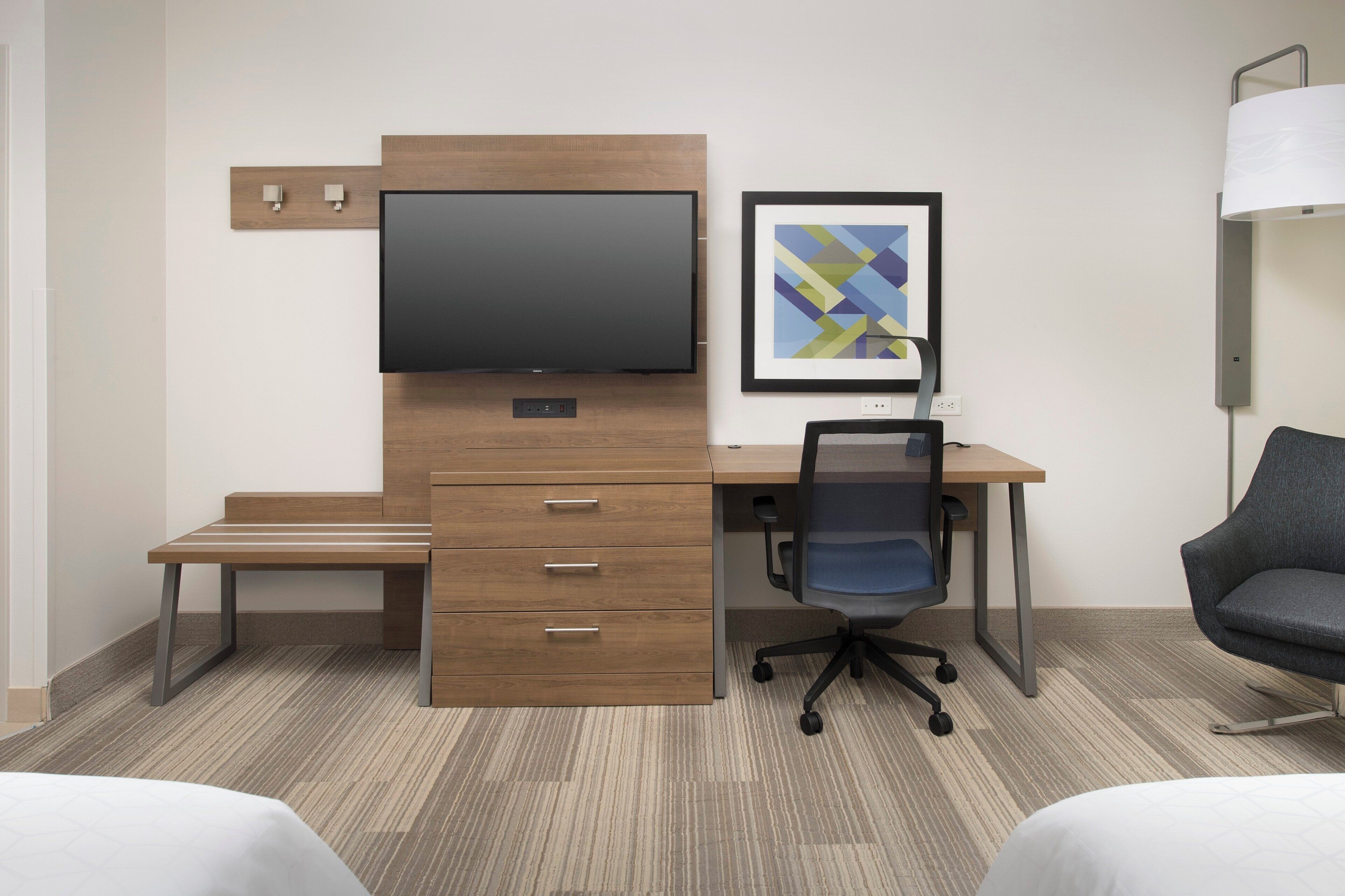 A hotel room with a wooden desk, a flat-screen TV, a framed abstract artwork, a desk lamp, a chair, and two beds.