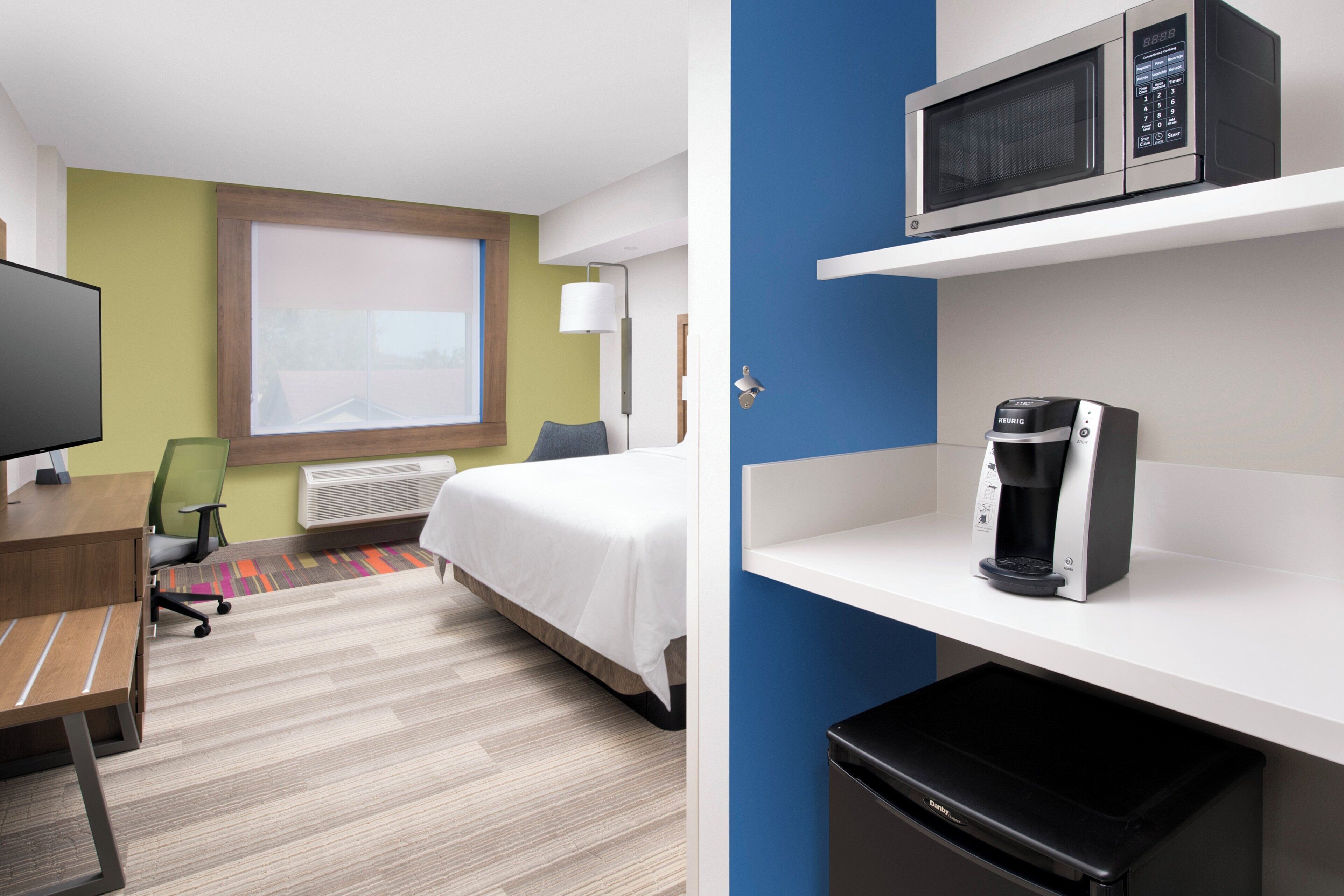 A hotel room with a bed, desk, chair, television, microwave, coffee maker, and a trash bin.