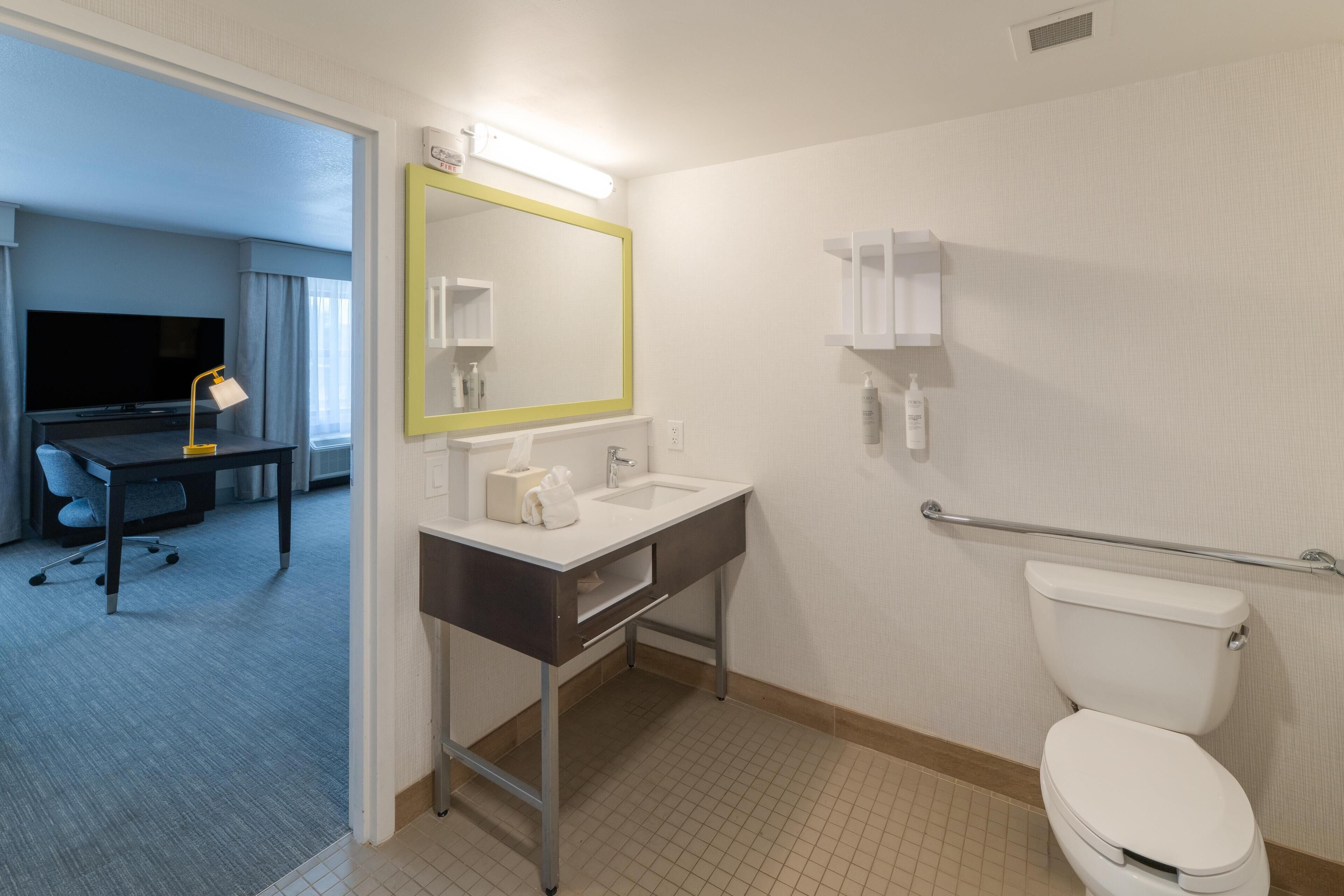 A hotel bathroom with a sink, mirror, toilet, and a handrail.
