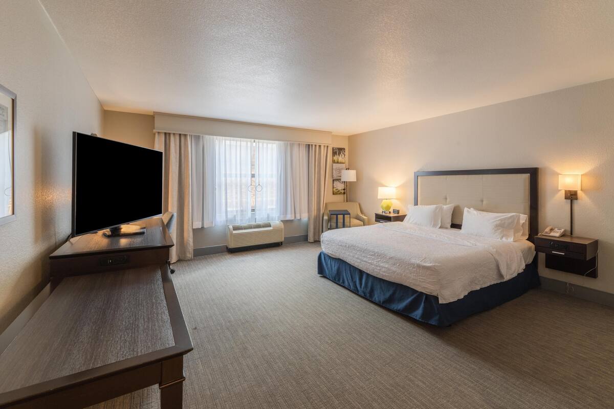 A hotel room with a large bed, a desk with a flat-screen TV, a bedside table with a lamp, and a window with curtains.
