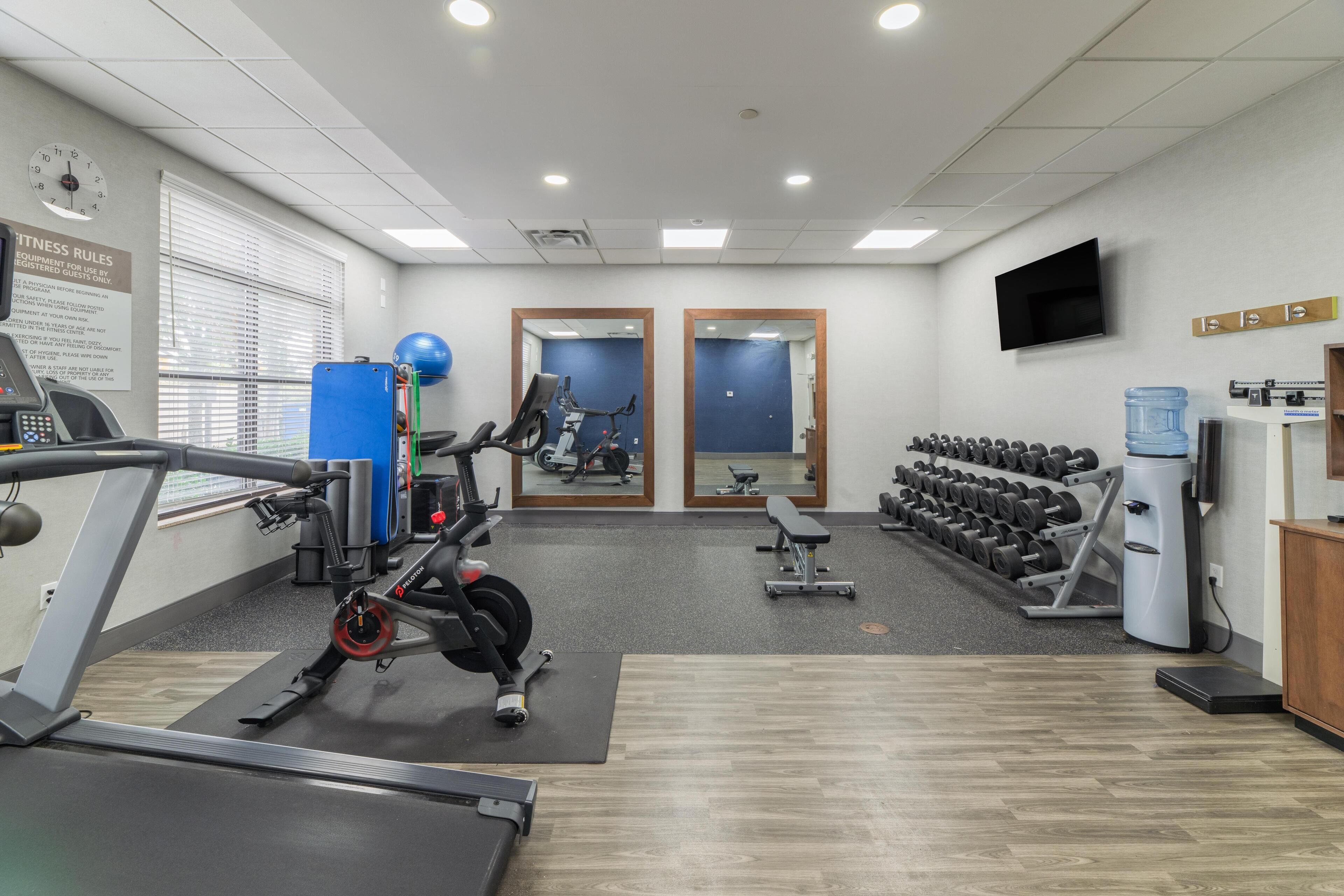 A gym with cardio equipment, weight machines, a bench, and a wall-mounted TV.