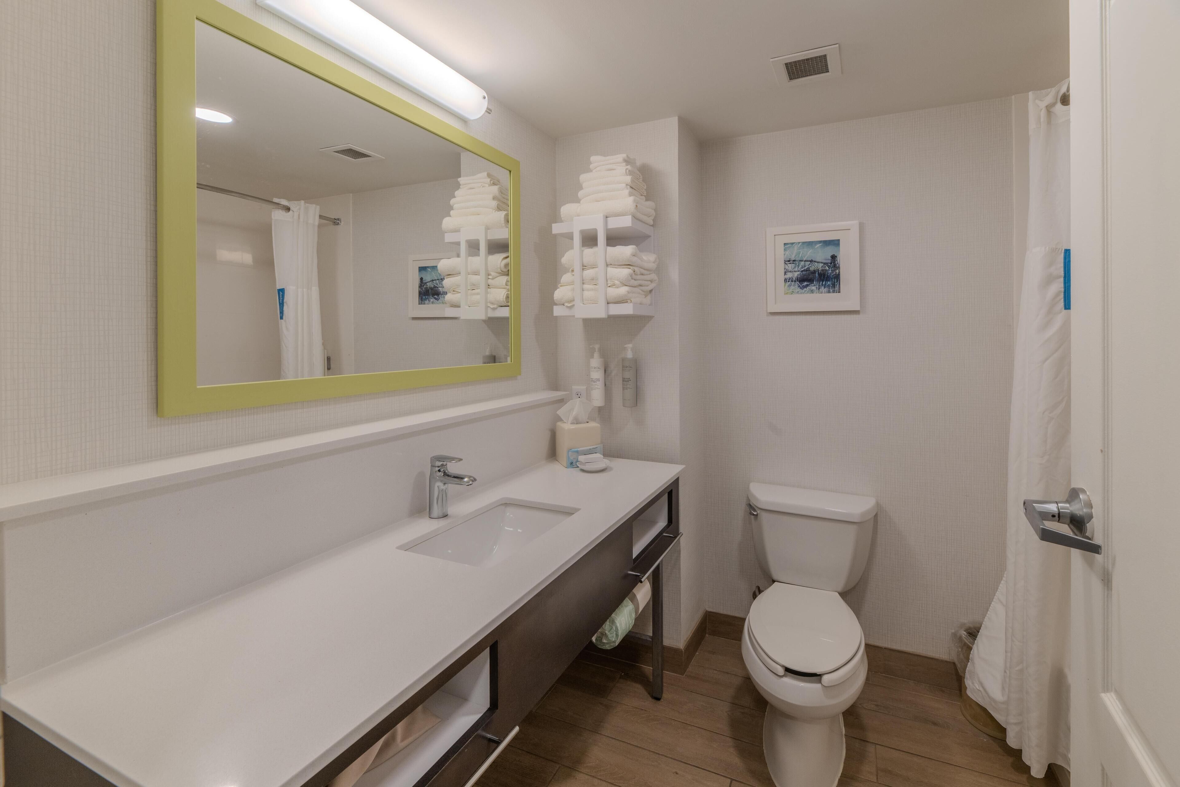A modern bathroom with a white sink, mirror, and toilet. There are shelves with folded towels and toiletries.