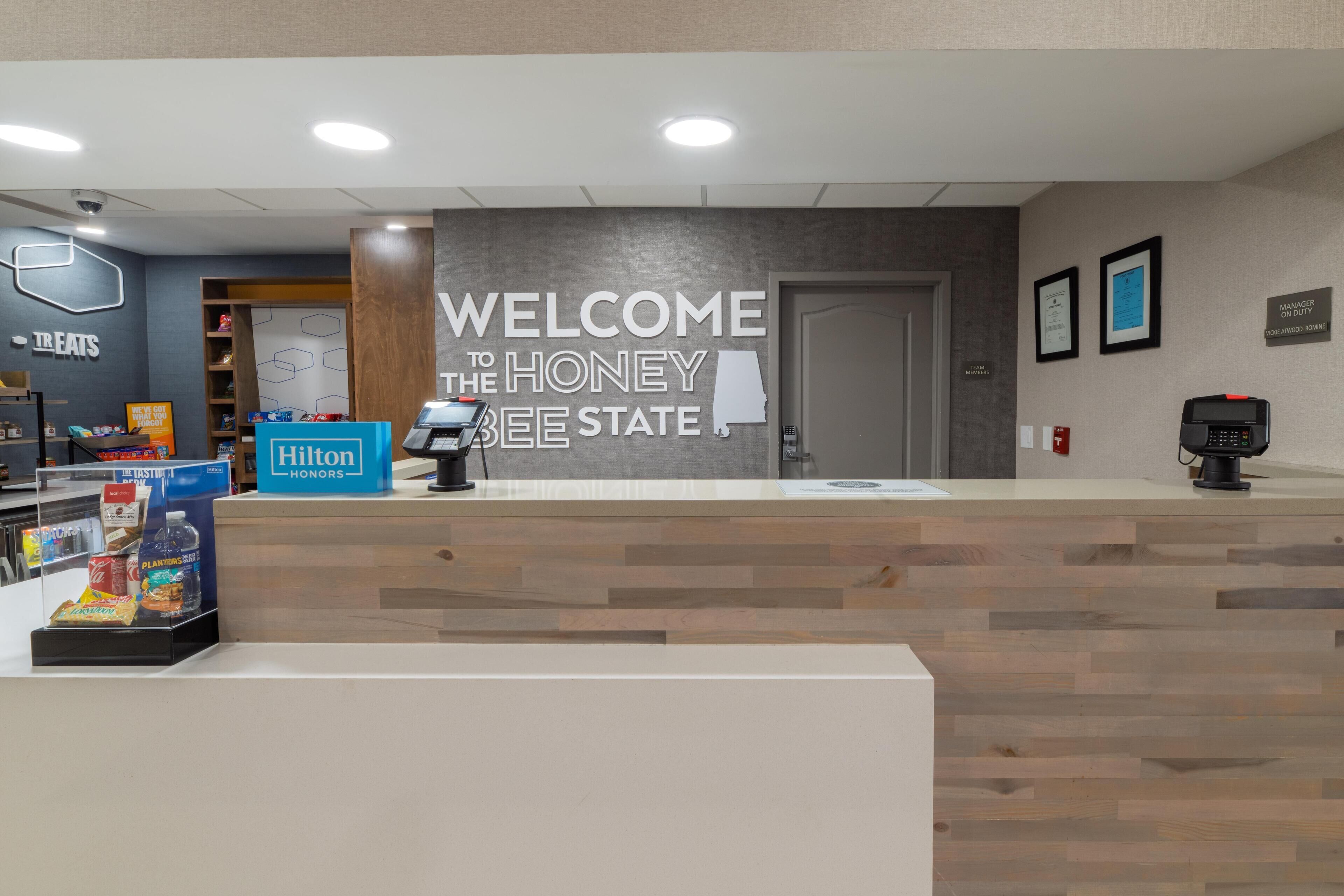 Uma área de recepção com uma placa 'Bem-vindo ao Honey Bee State', um sinal de Hilton Honors e um balcão com telefone e um pequeno expositor.