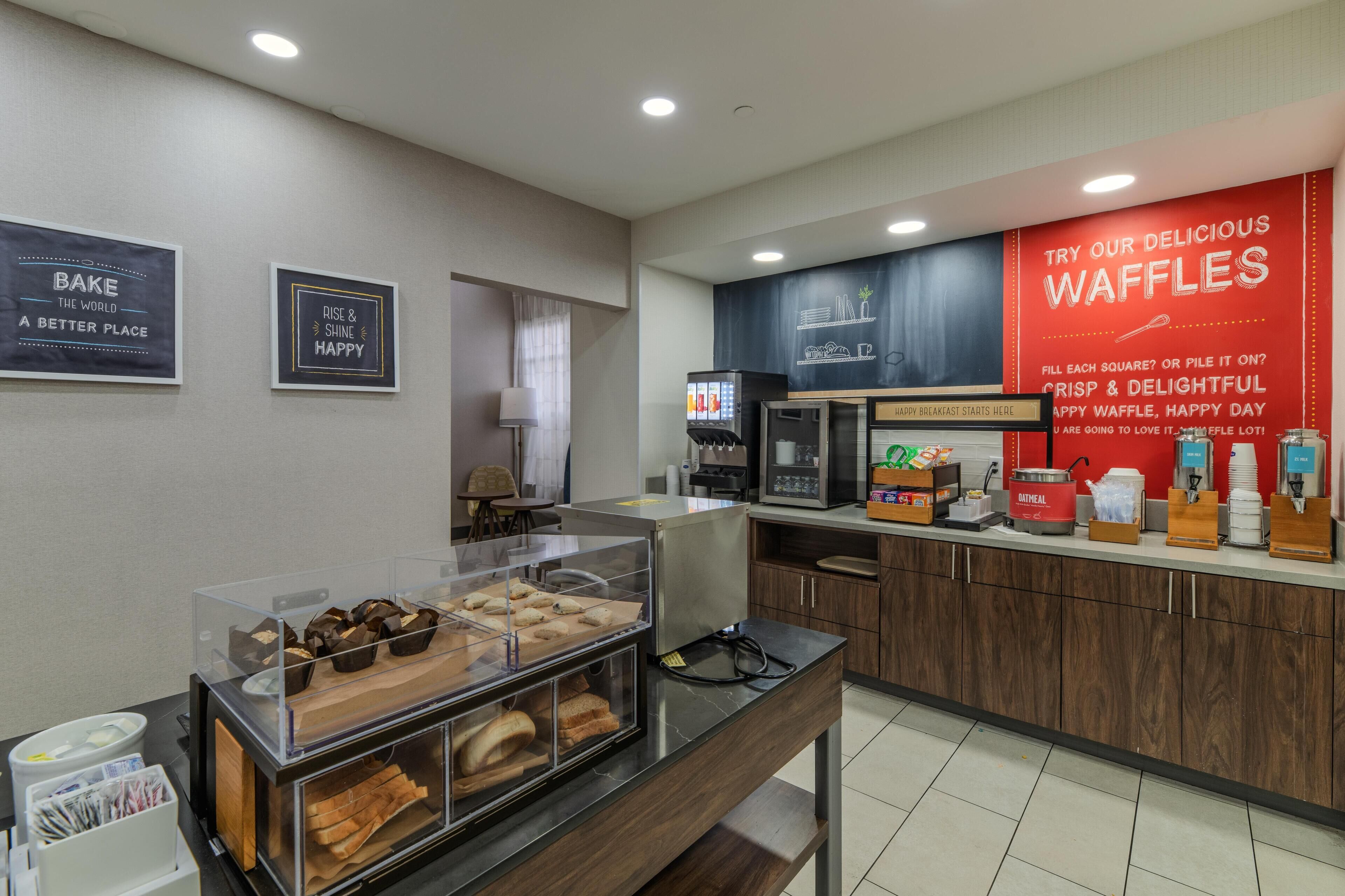 Un área de desayuno con una estación de gofres, una cafetera y una vitrina con productos de repostería.