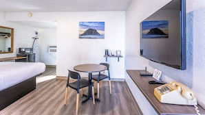 A hotel room with a bed, a desk with a telephone, a chair, a small table, and two paintings on the wall. - Coastal Breeze Inn (Morro Bay)
