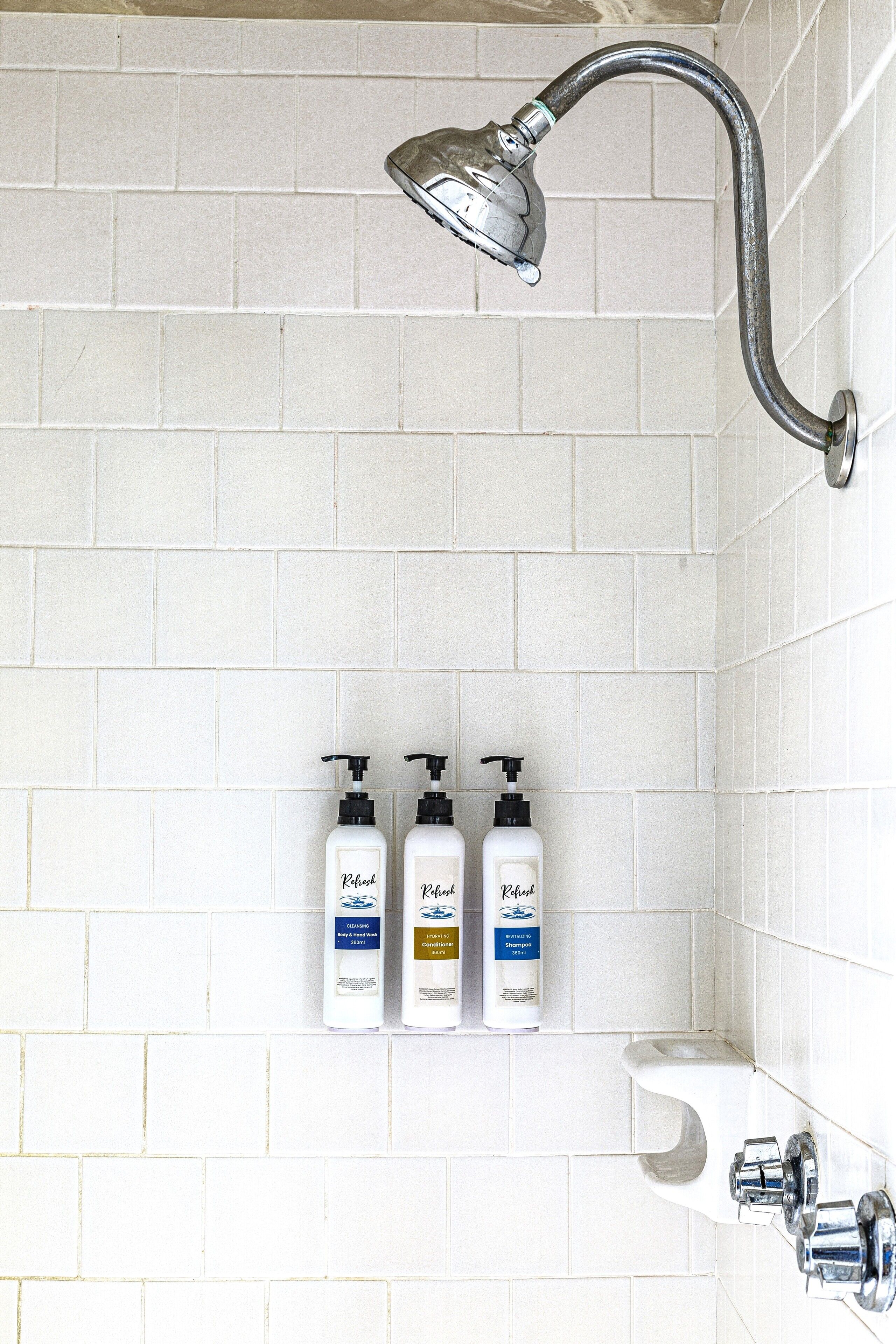 Um chuveiro com um chuveiro manual, parede revestida de azulejos brancos e três frascos de shampoo em uma prateleira.