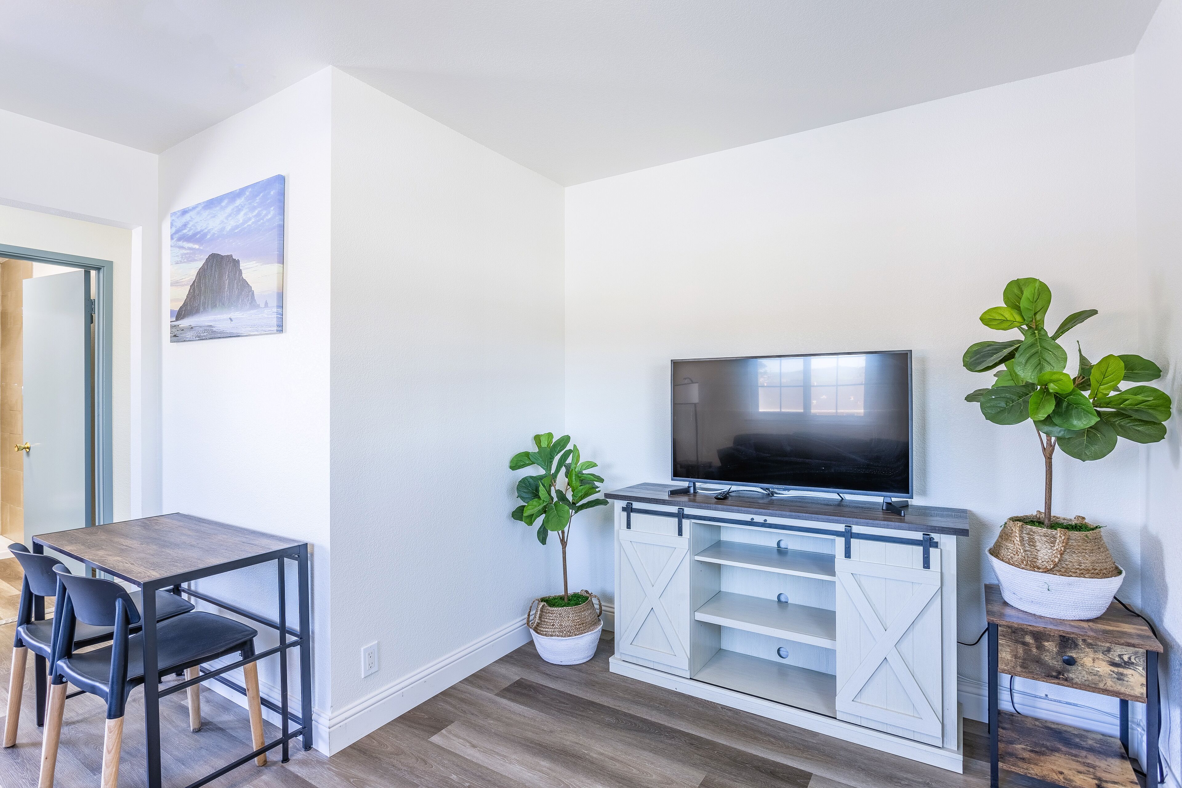 A modern living room with a wooden dining table, two chairs, a flat-screen TV, and two potted plants.