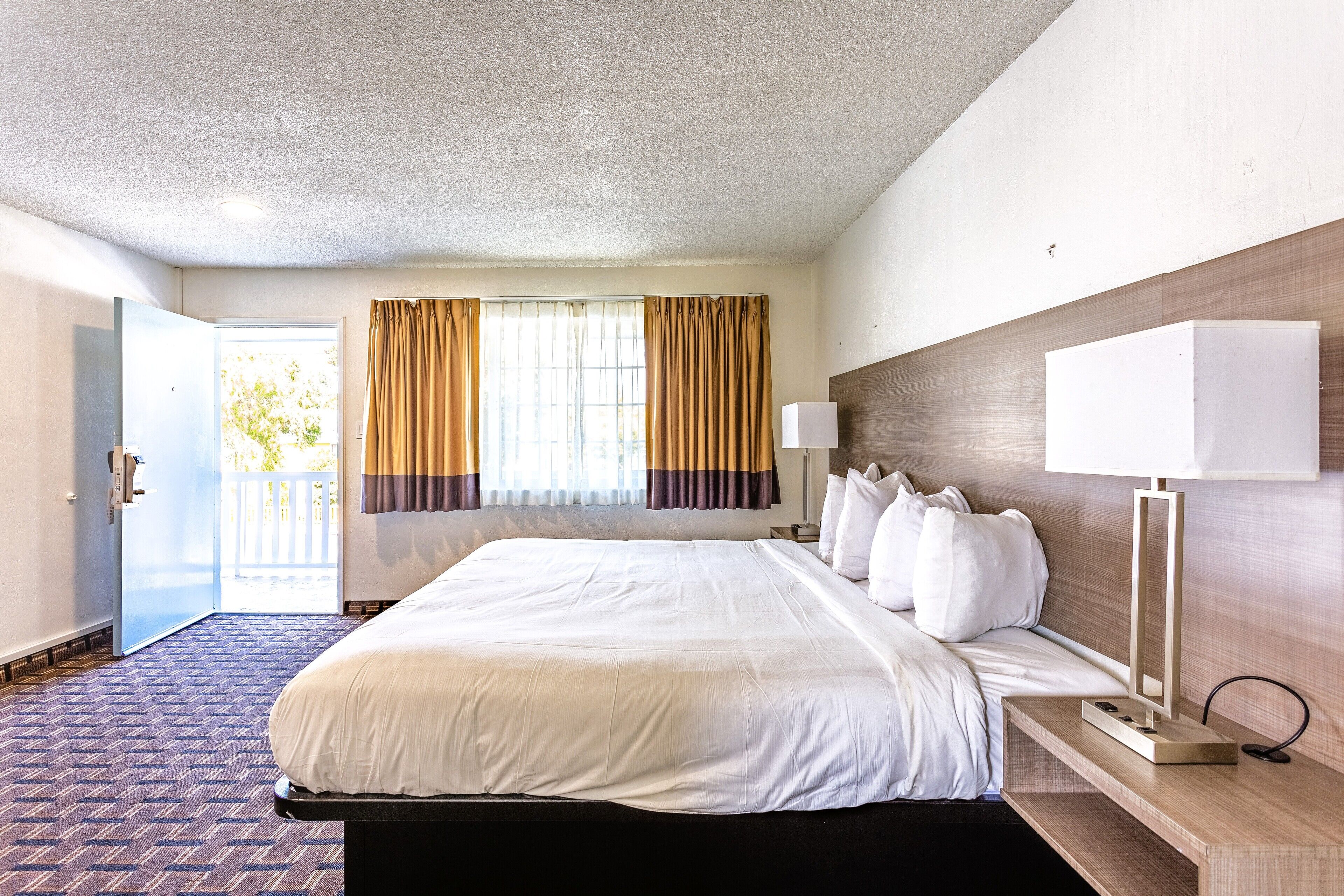 A hotel room with a large bed, a bedside table with a lamp, a door leading to a balcony, and curtains on the windows.