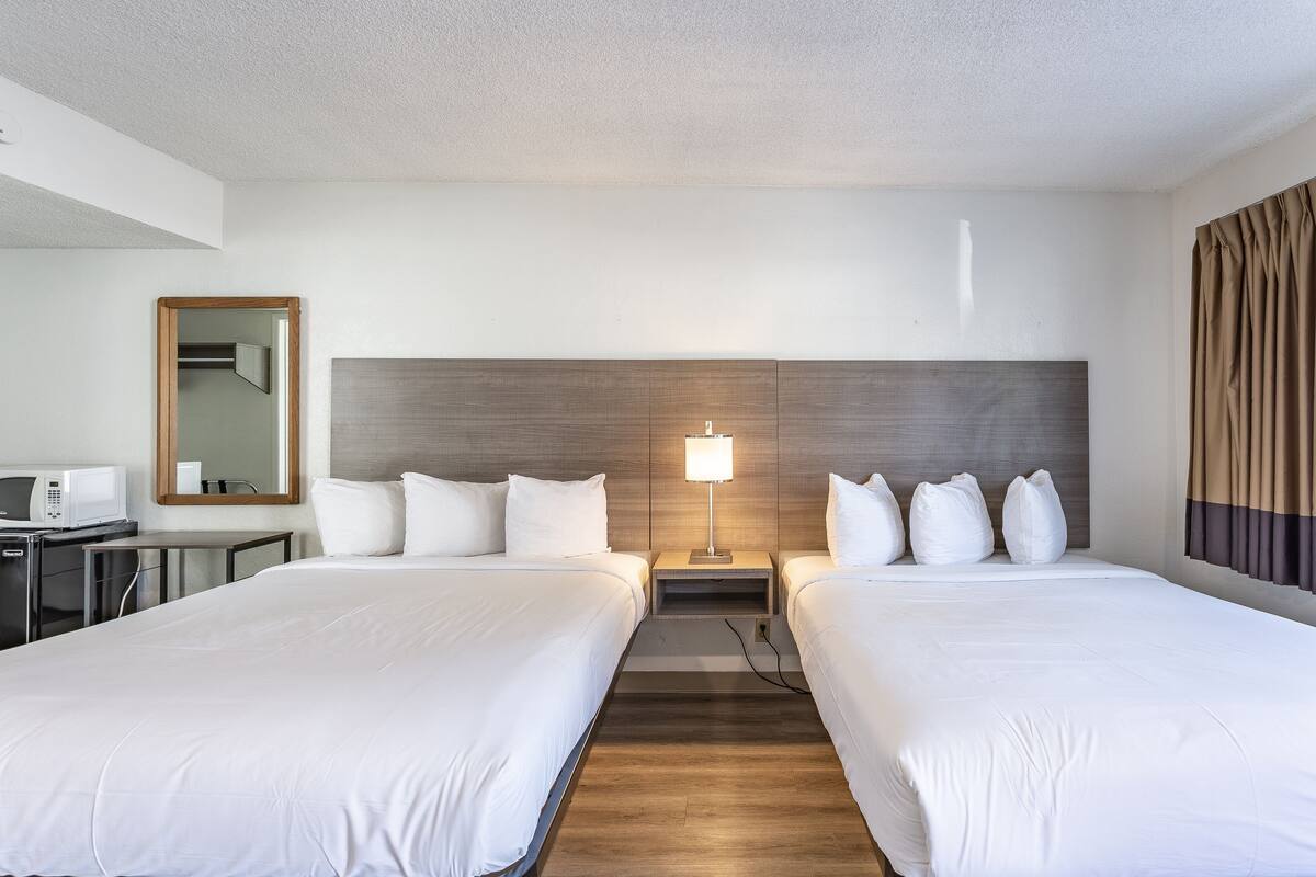 Una camera d'albergo con due letti, un comodino con una lampada, uno specchio e una scrivania con un forno a microonde.