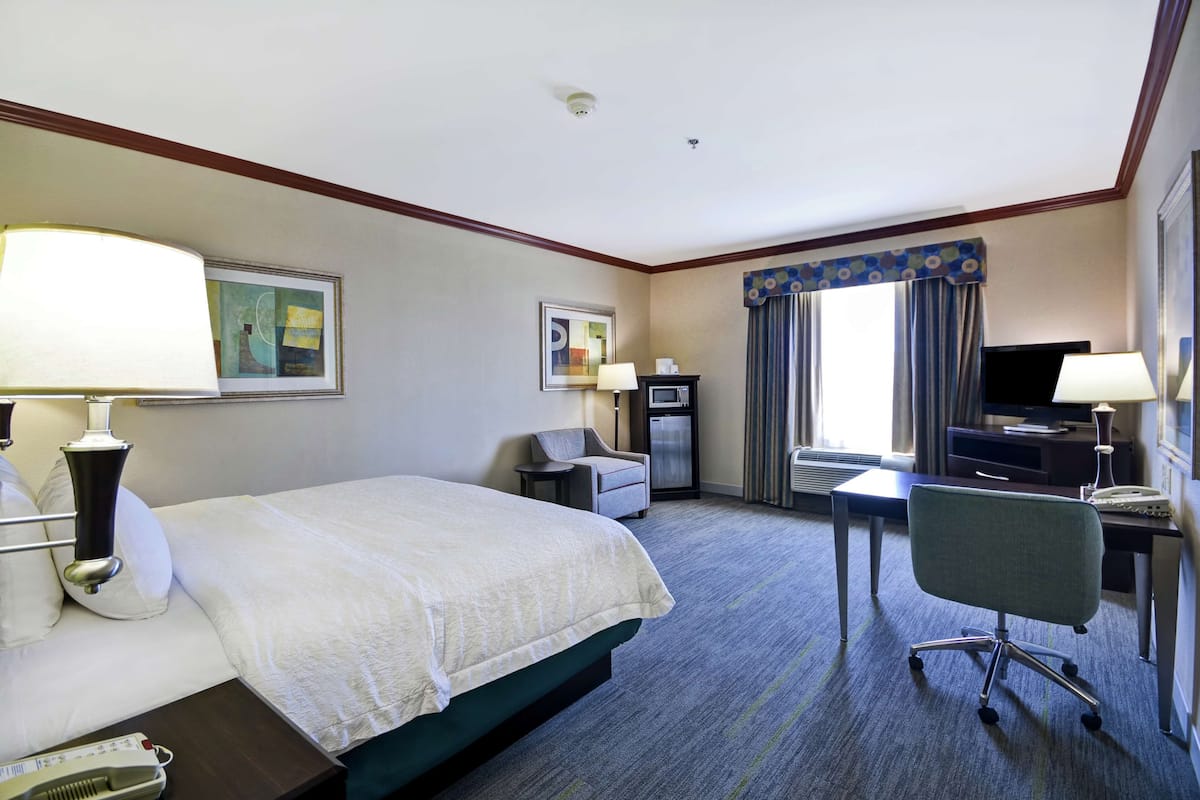 A hotel room with a bed, desk, chair, TV, and artwork on the walls.