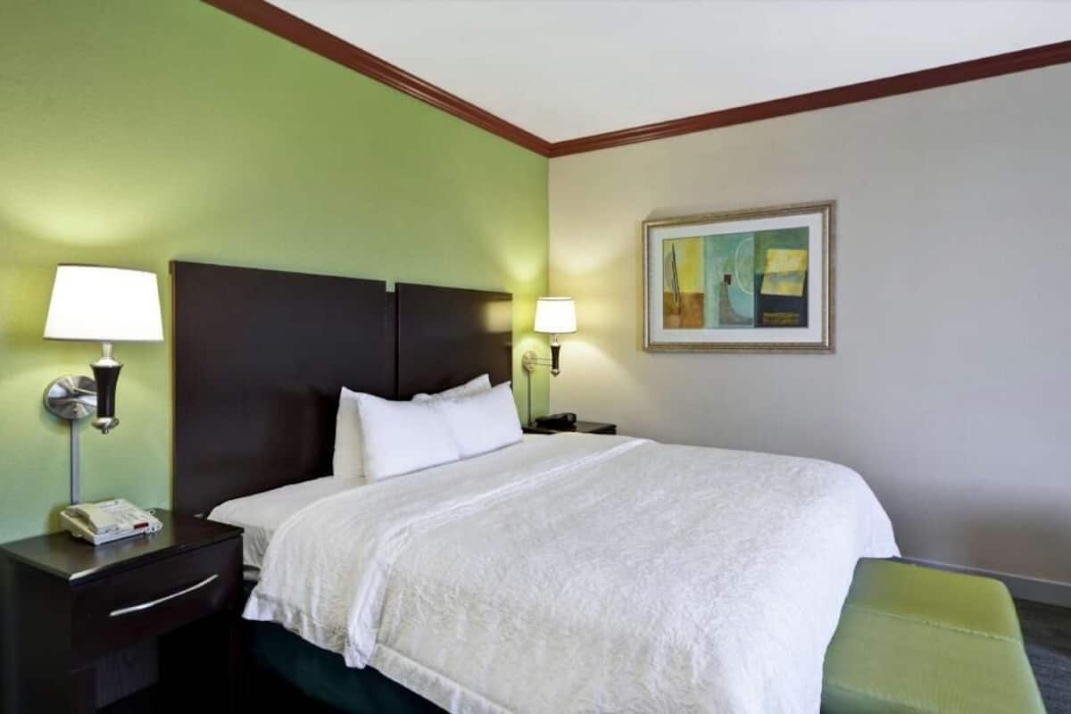 A hotel room with a bed, two bedside tables with lamps, a painting on the wall, and a green chair.