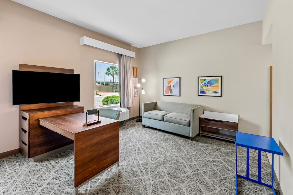 A hotel room with a wooden desk, a flat-screen TV, a gray sofa, and two framed artworks.