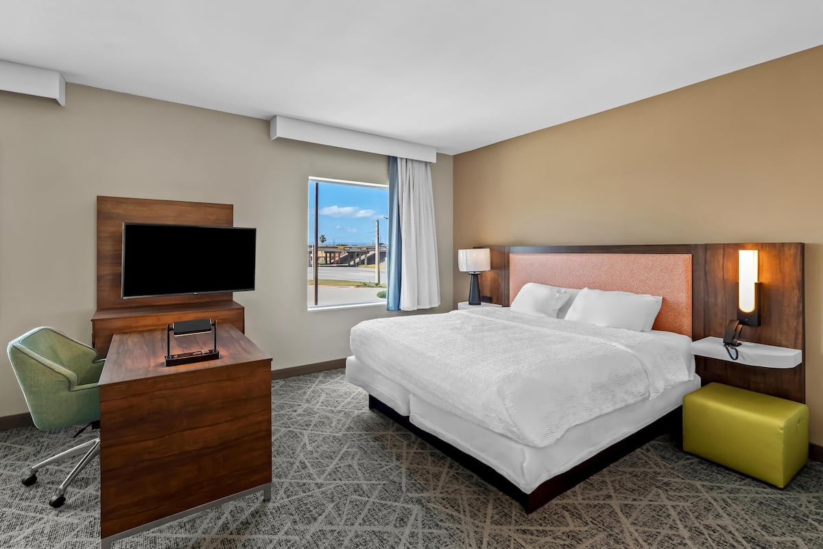 A hotel room with a large bed, a desk with a flat-screen TV, a green chair, and a window with a view of a bridge.