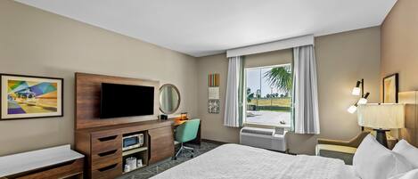 A hotel room with a large bed, a desk with a chair, a television, a window with curtains, and a view of the outdoors.