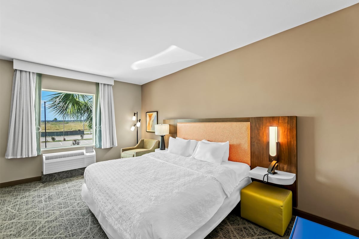A hotel room with a large bed, a bedside table with a lamp, a chair, and a view of the outdoors.
