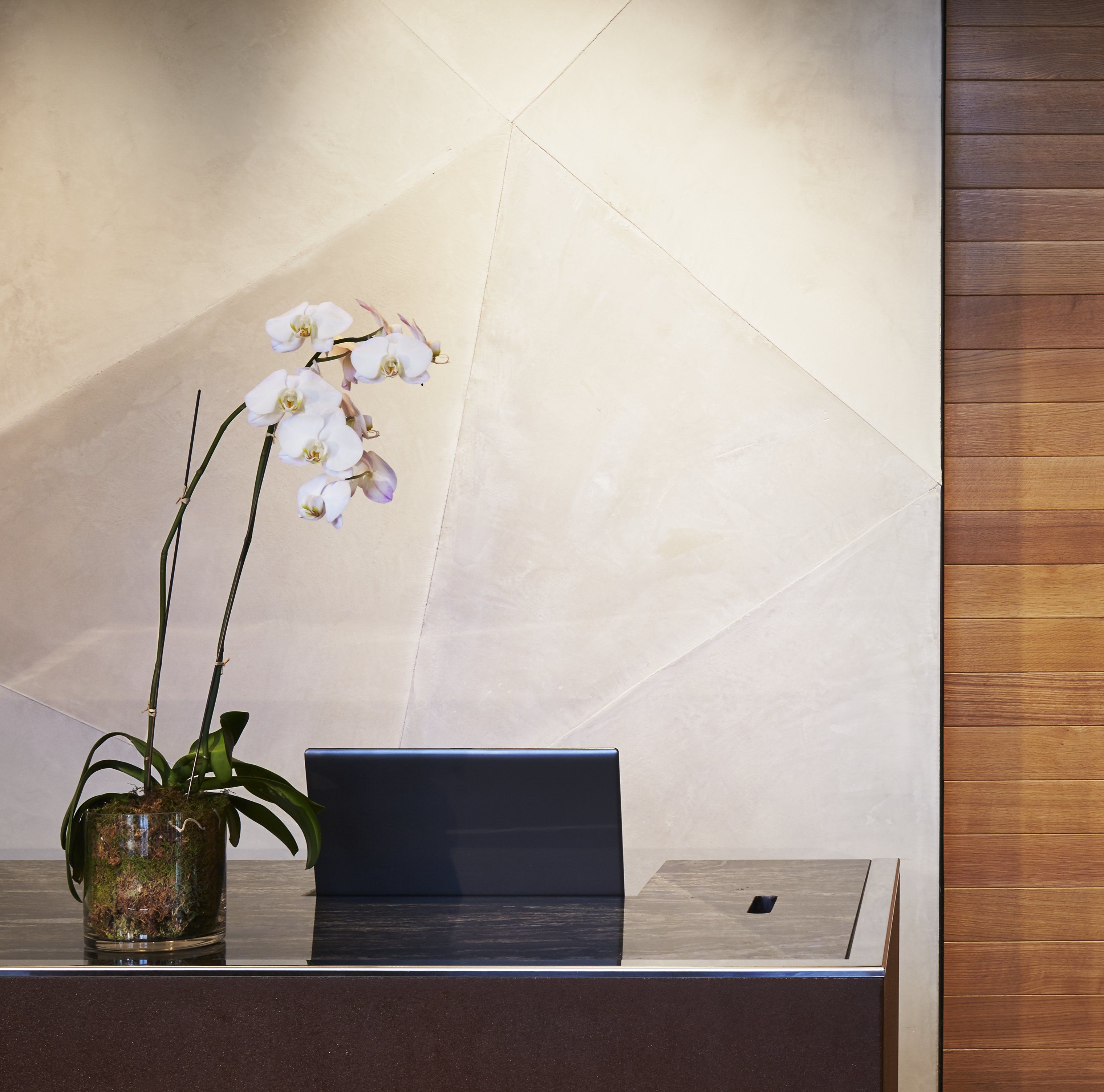 A reception desk with a laptop, a vase with orchids, and a wooden panel wall.