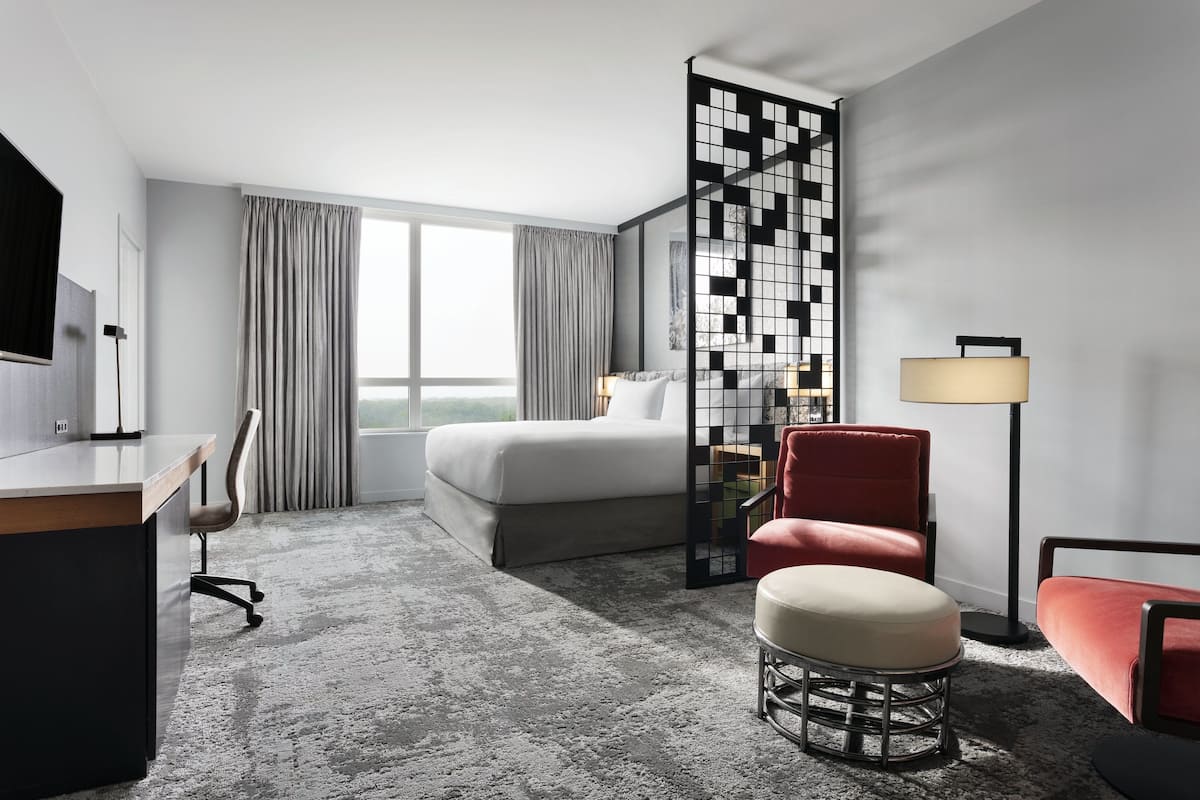 A hotel room with a bed, desk, chair, ottoman, floor lamp, and a decorative partition.