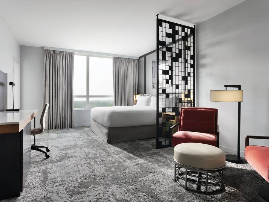A hotel room with a bed, desk, chair, ottoman, floor lamp, and a decorative partition.