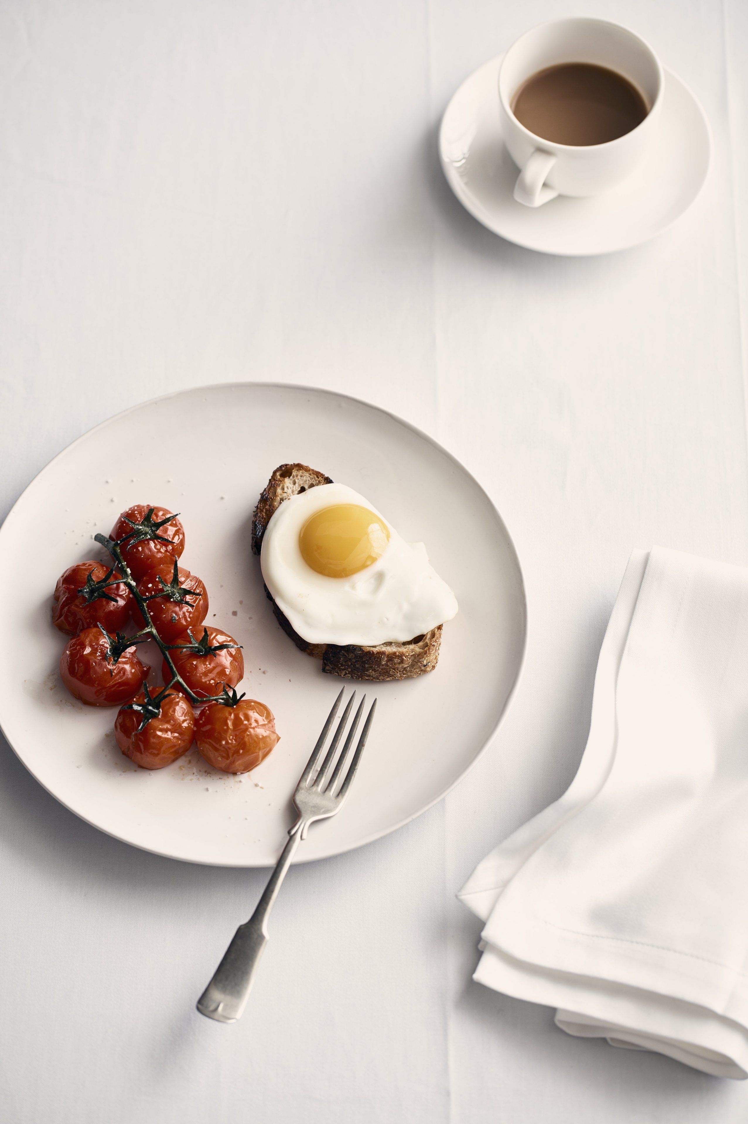 A plate with a fried egg on toast and cherry tomatoes, a fork, and a cup of coffee on a white table.