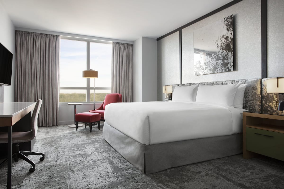 A modern hotel room with a large bed, a desk, a chair, and a view of the outdoors.