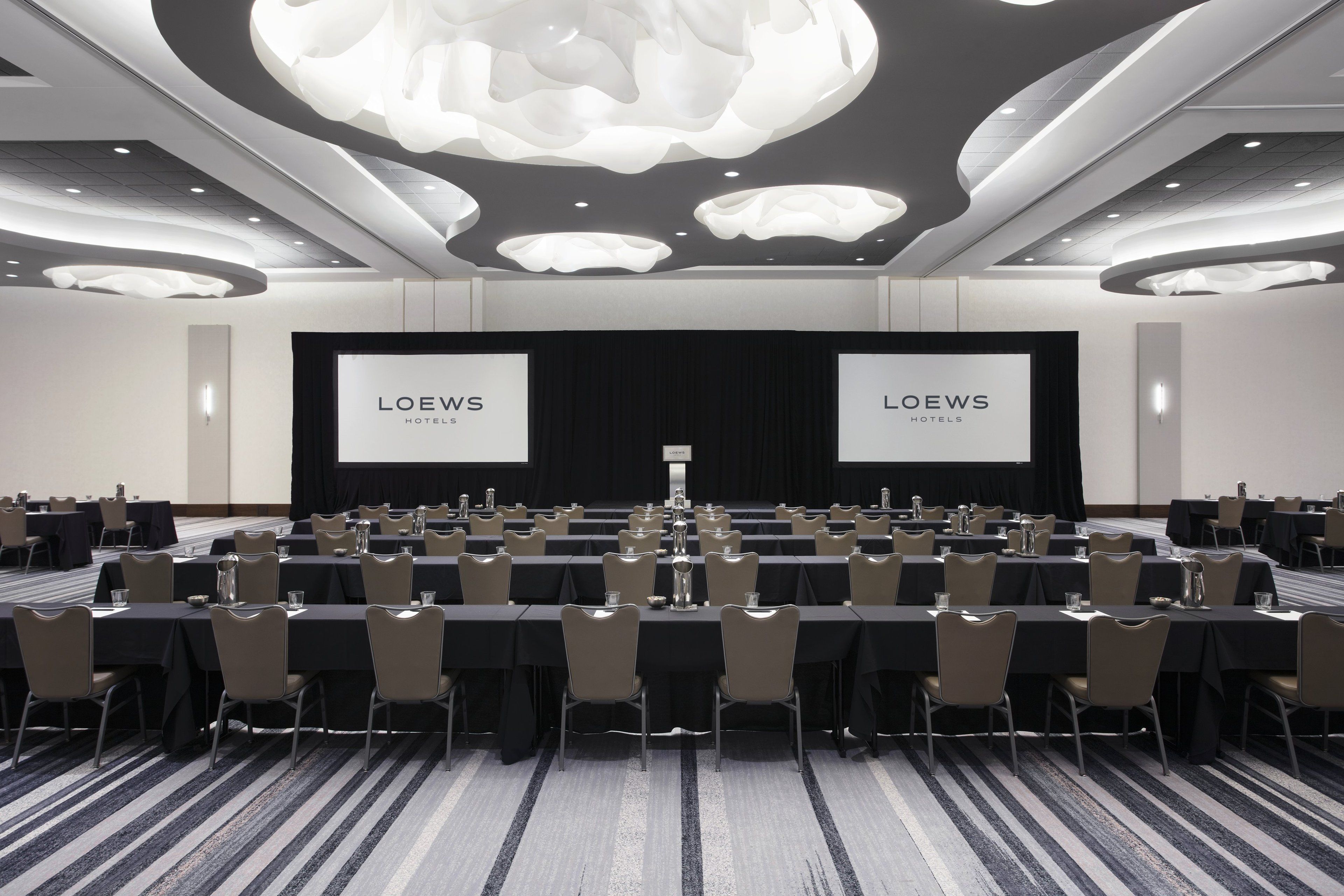 Une salle de conférence dotée de plusieurs tables et chaises, d’un grand écran affichant le nom « LOEWS HOTELS », et d’un design de plafond moderne.