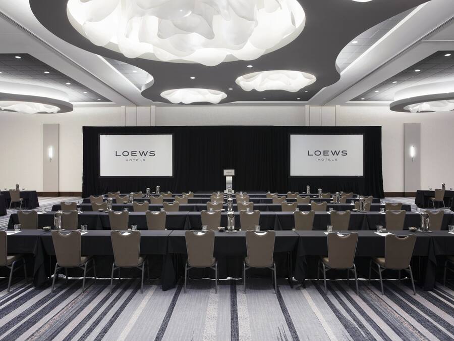 Une salle de conférence dotée de plusieurs tables et chaises, d’un grand écran affichant le nom « LOEWS HOTELS », et d’un design de plafond moderne.