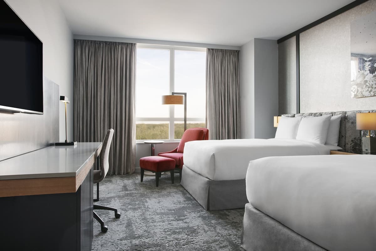 A hotel room with a desk, two beds, a chair, and a large window with curtains.