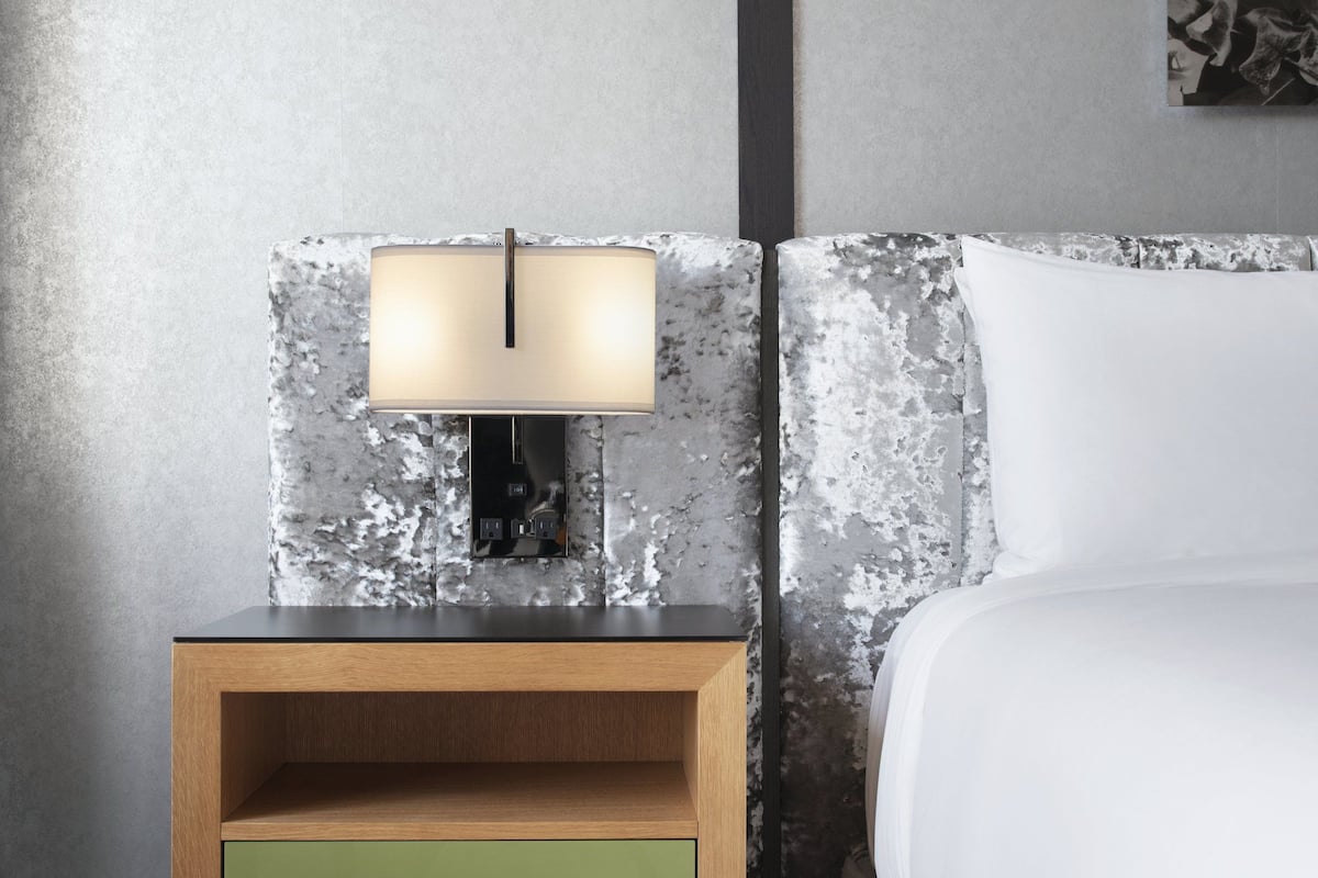 A bedroom with a wooden nightstand, a green drawer, a wall-mounted lamp, and a bed with white bedding.